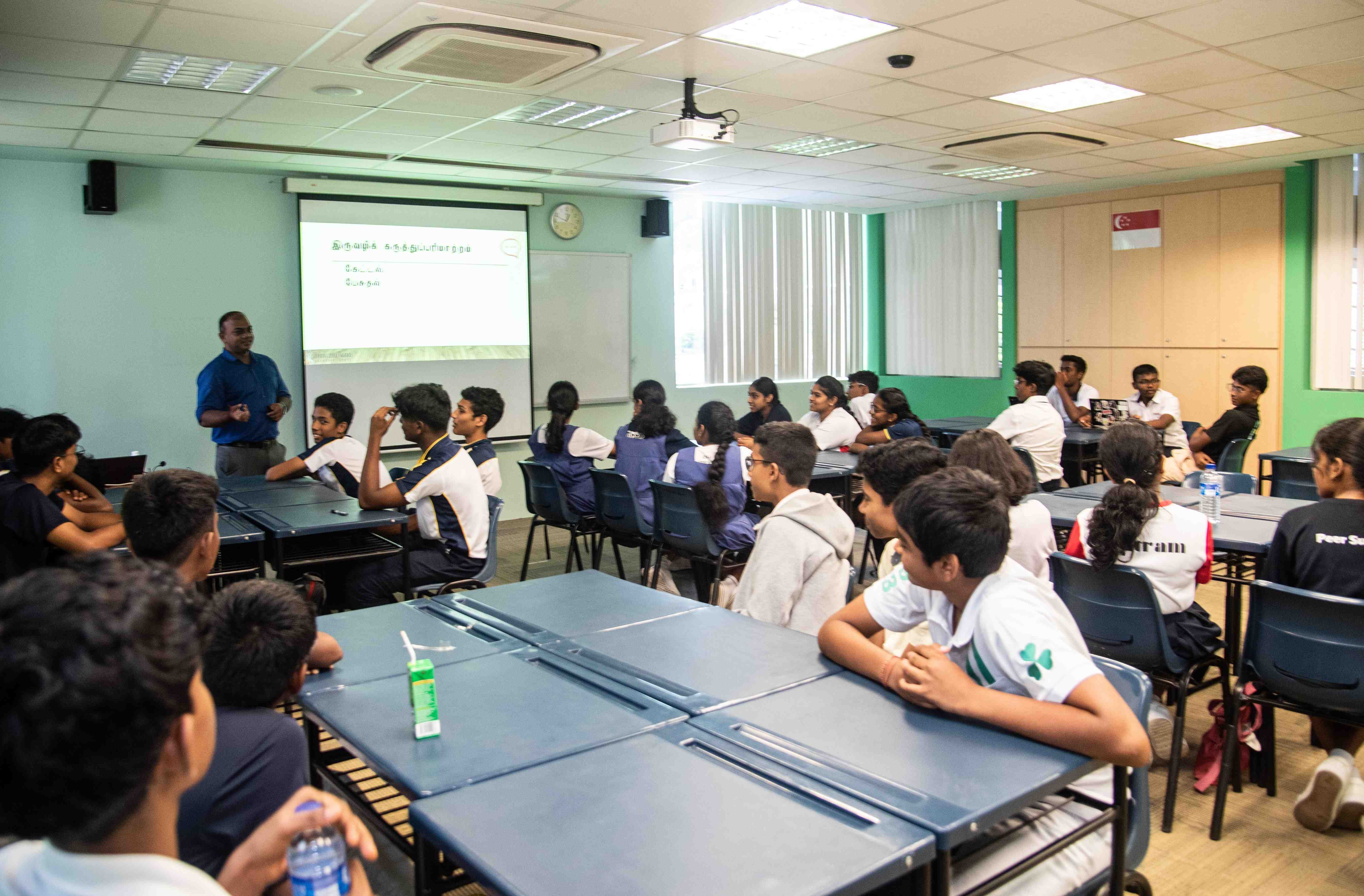 Teacher stands by projector screen addressing seated students at tables in a classroom.