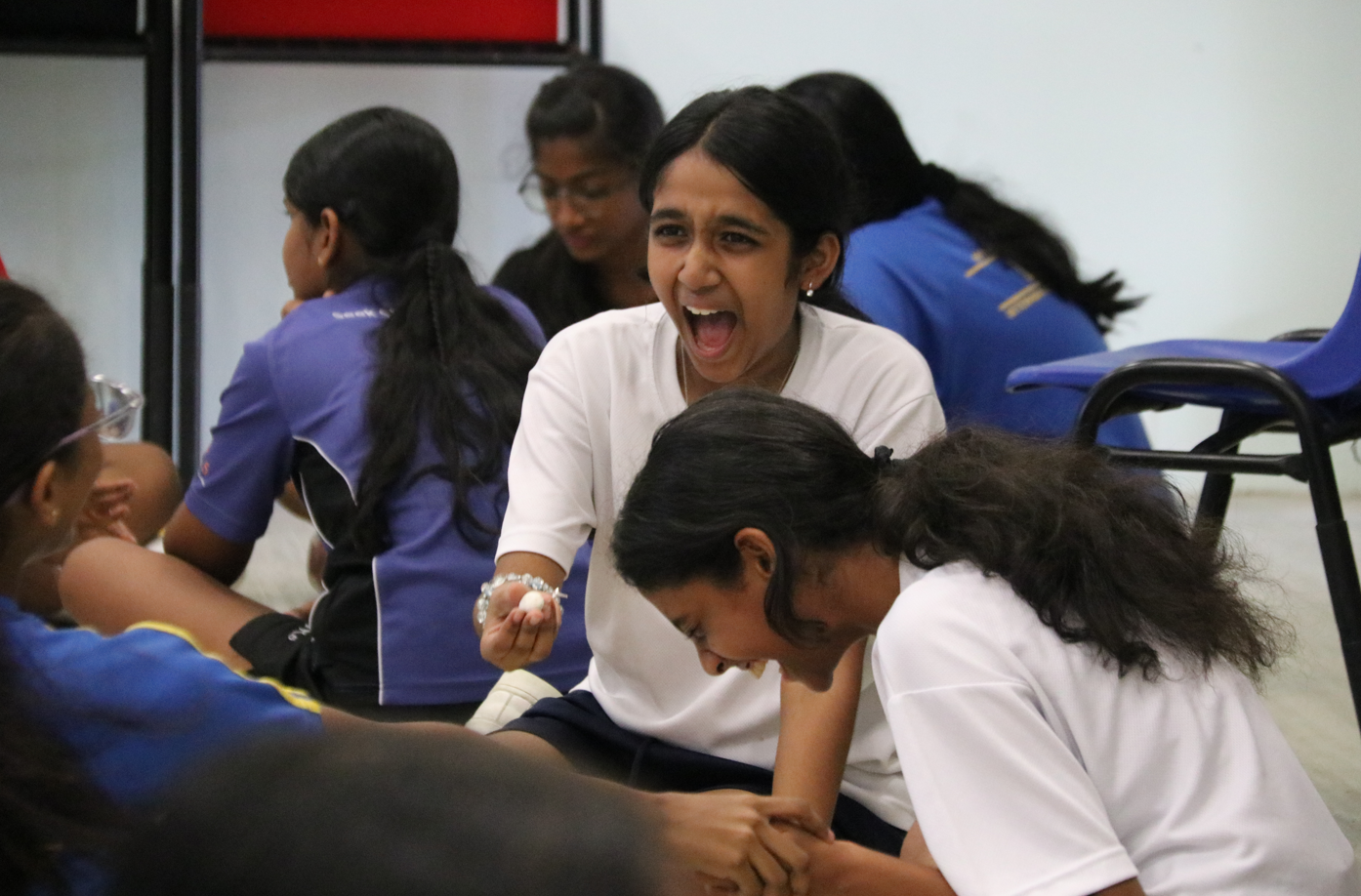 Girl in white shirt shouts while holding a small object as another girl laughs; others sit nearby.