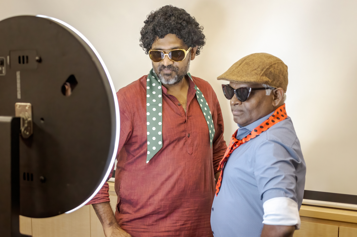 Two men wearing sunglasses and polka-dot scarves pose beside a large ring light.
