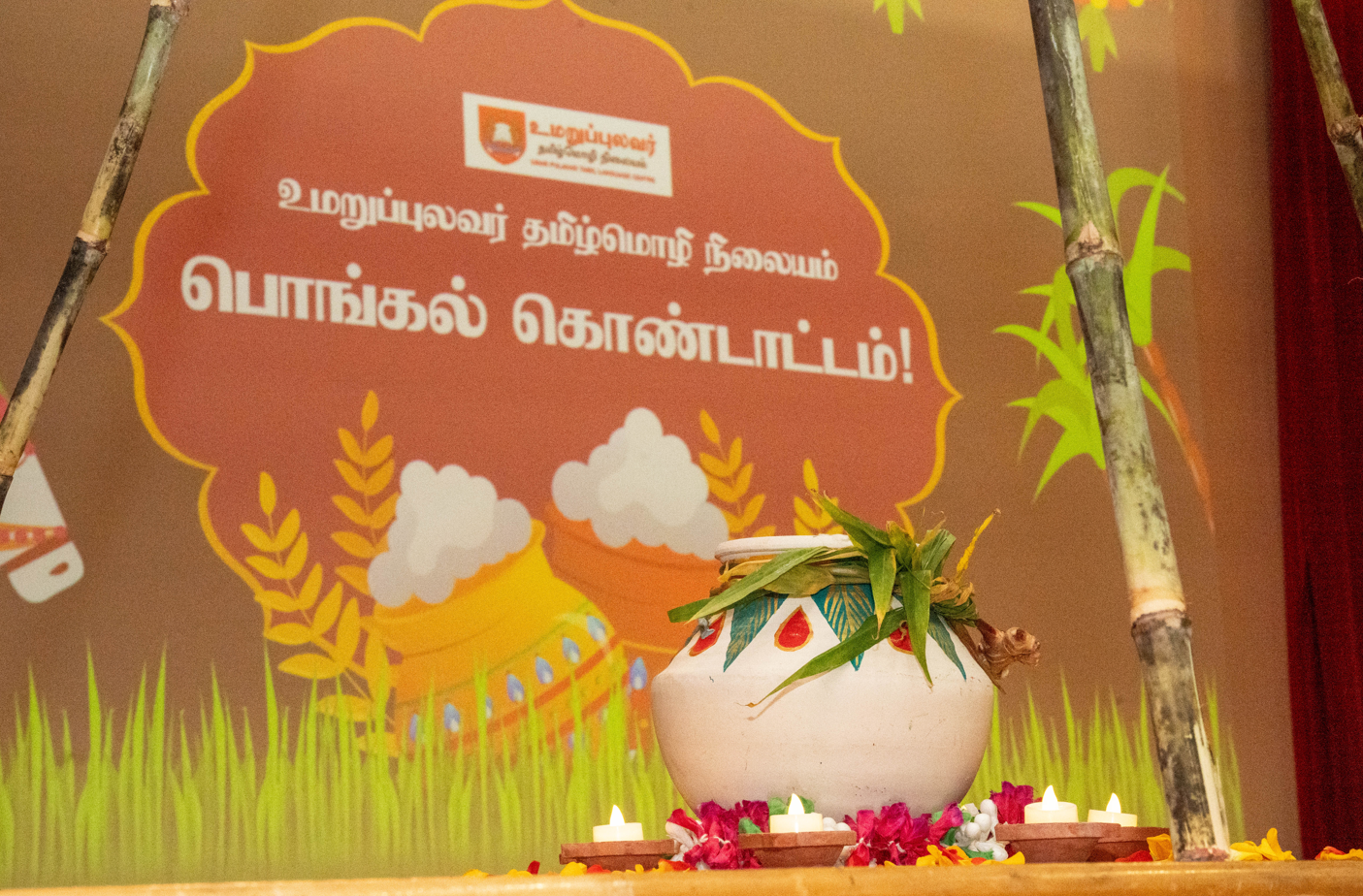 Decorated white clay pot with leaves and lit oil lamps in front of a Tamil festival banner and sugarcane stalks.