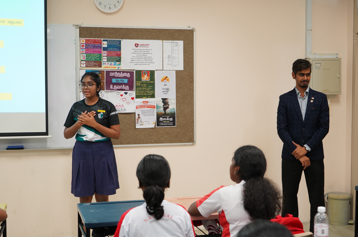 Student speaks in front of bulletin board while a man stands nearby; classmates sit facing them in a classroom.