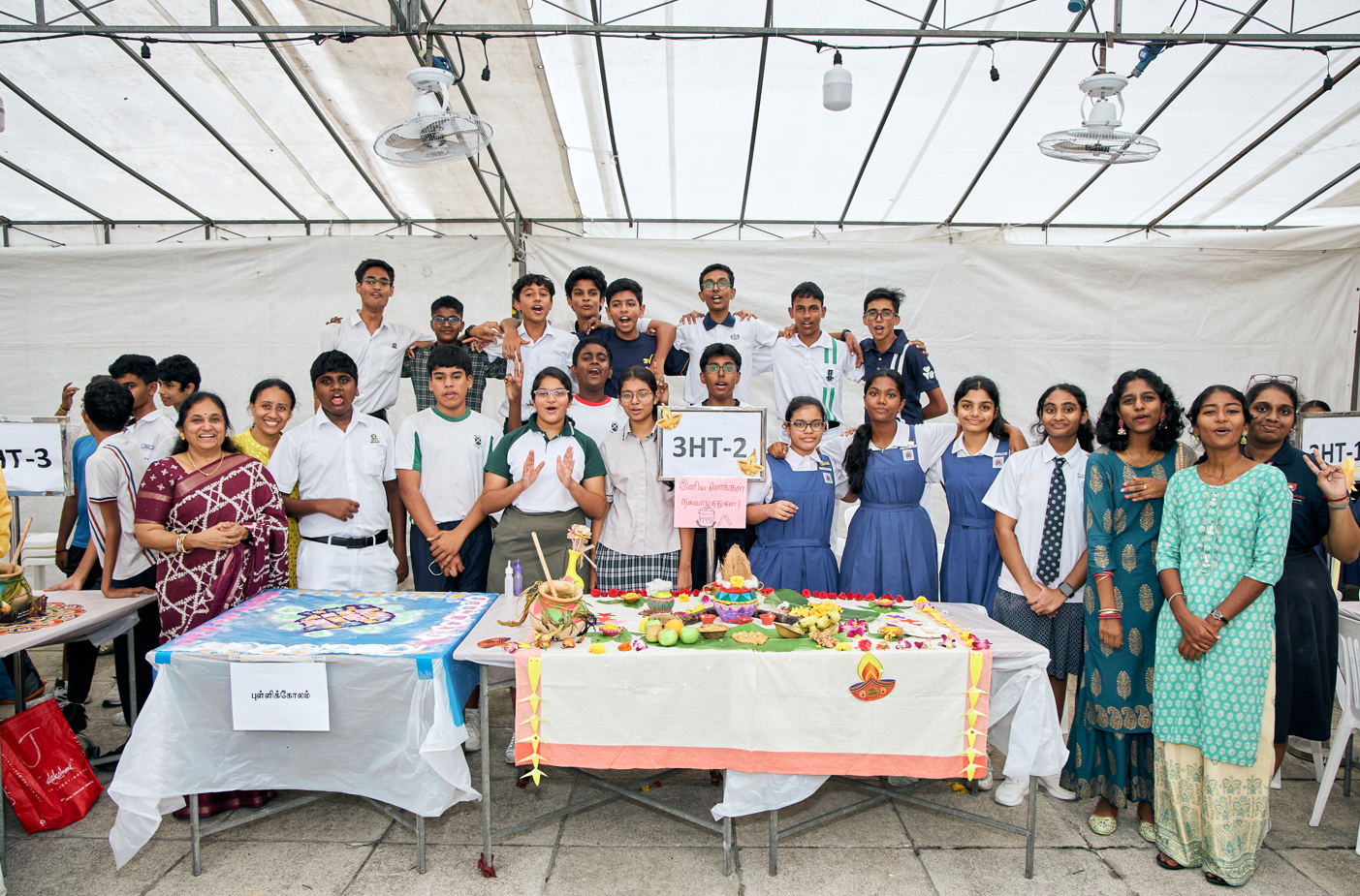 Group of students and teachers posing behind decorated tables at a school fair, holding a 3HT-2 sign