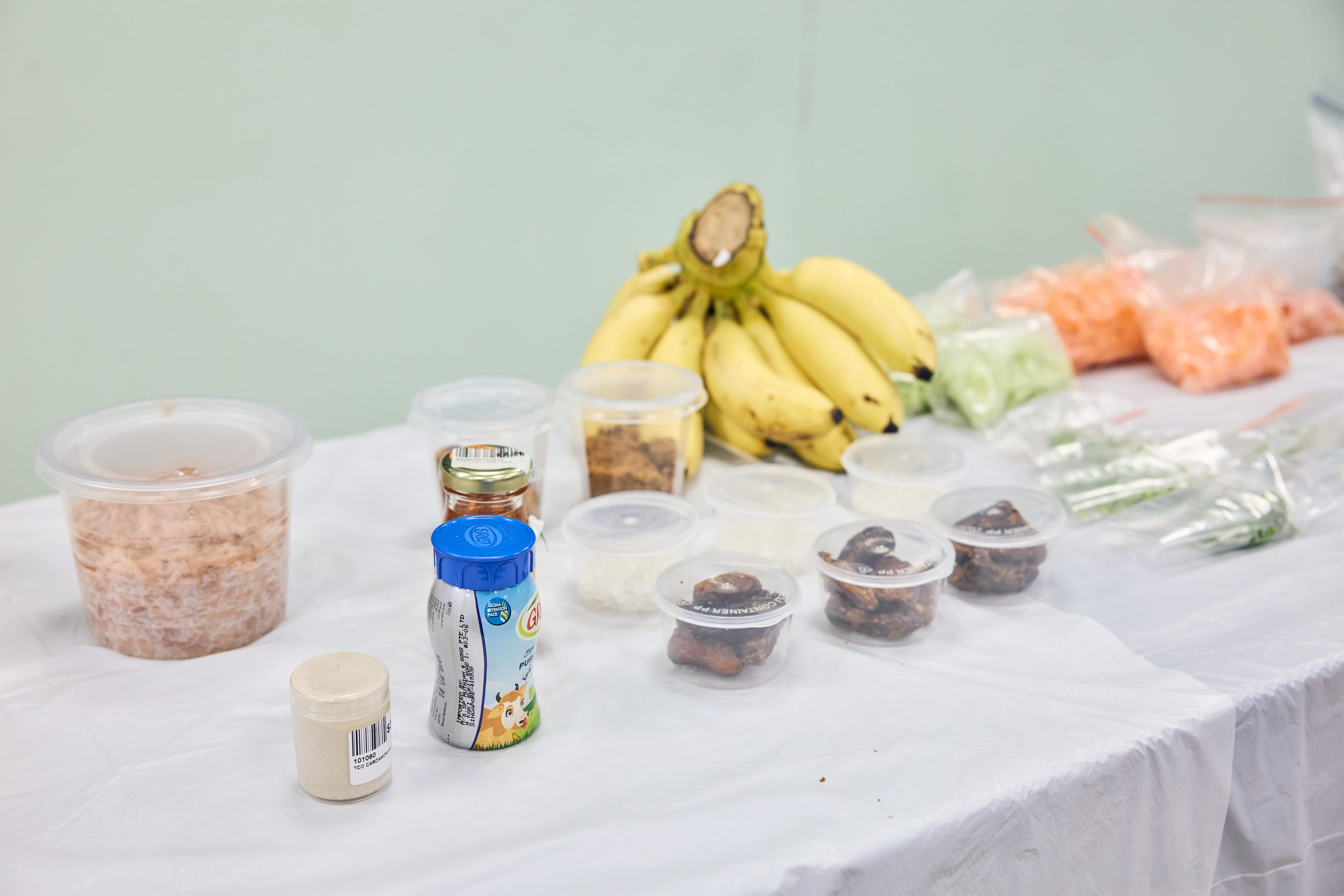 Table with bananas, small food containers, a yogurt drink, and bagged vegetables on a white cloth.