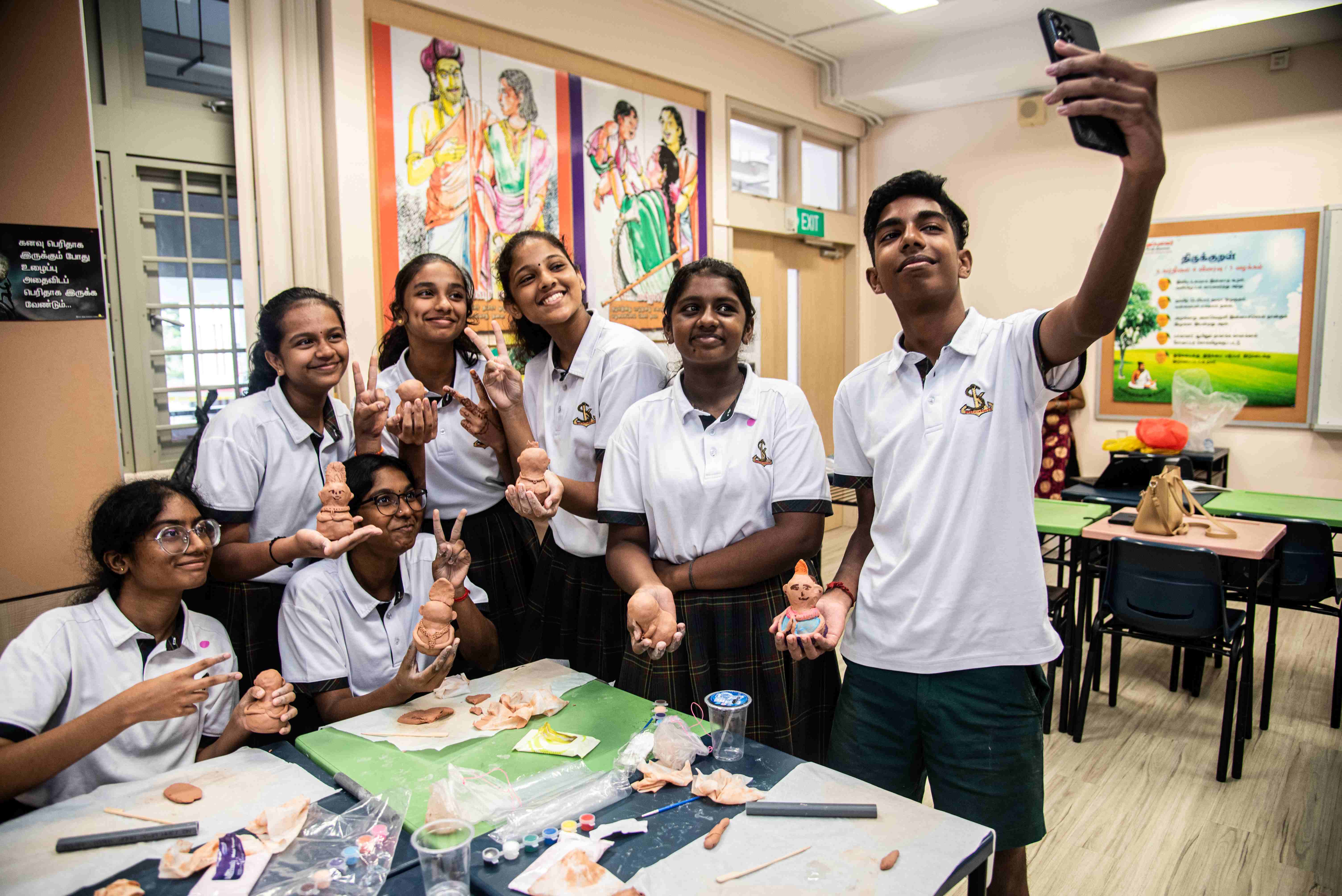 Group of students in uniforms pose in a classroom, holding clay figures while one takes a selfie.