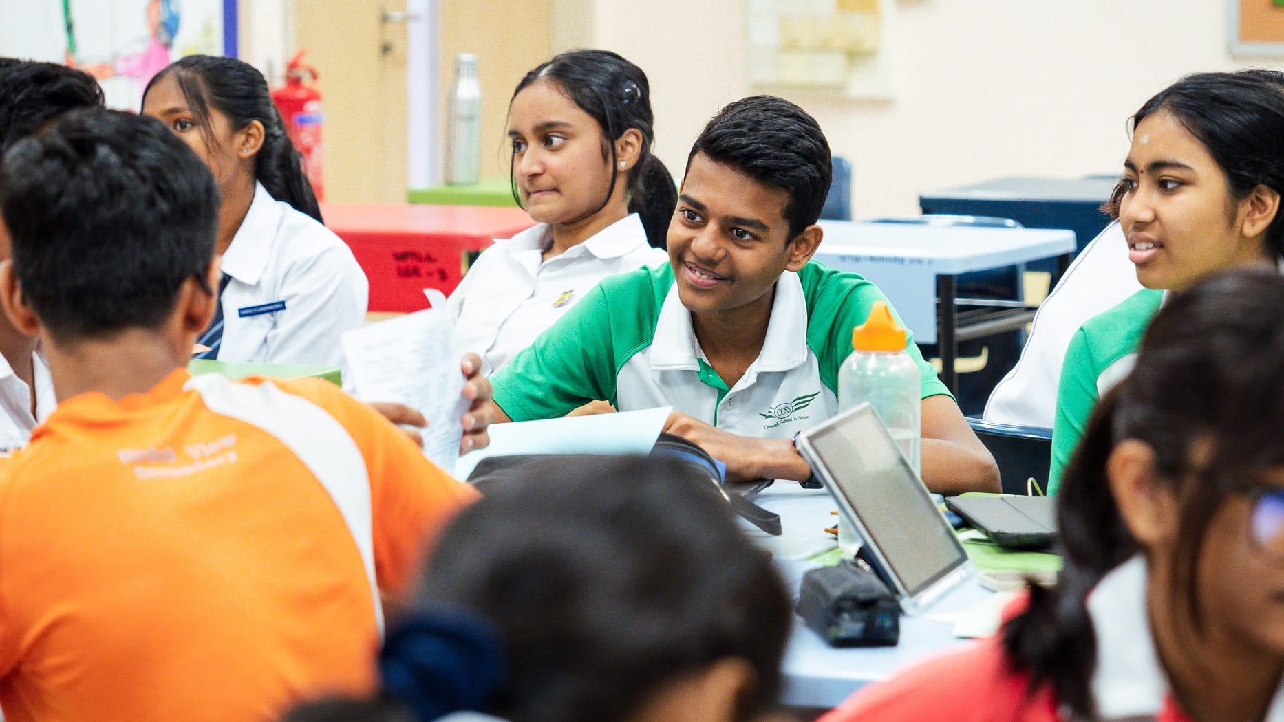 Students in uniforms sit around a table in class, one boy smiles while talking to others.