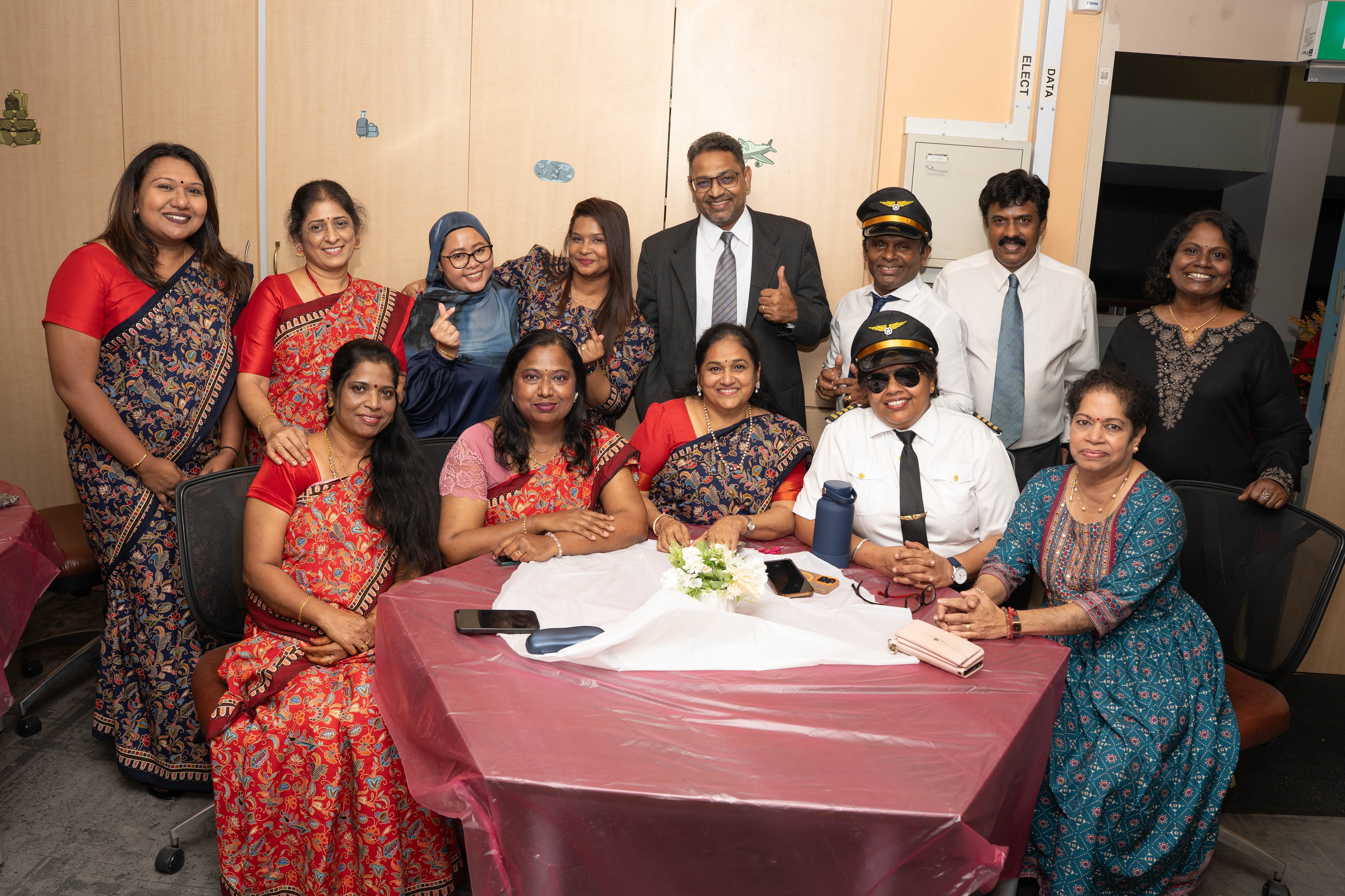 A group of about 13 smiling people posing together, some in traditional Indian attire, one wearing a pilot uniform and cap.