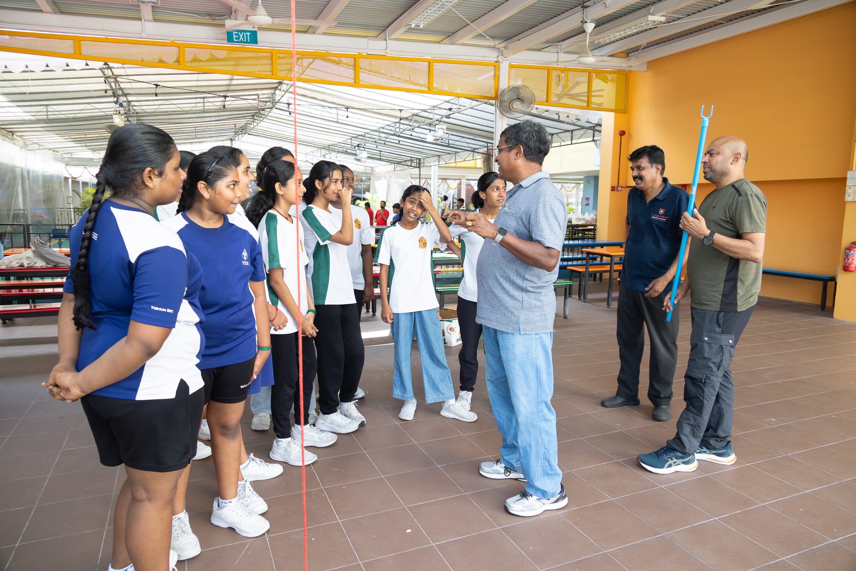 Group of students in sports uniforms listen to a man speaking in a school hall while another holds a pole.