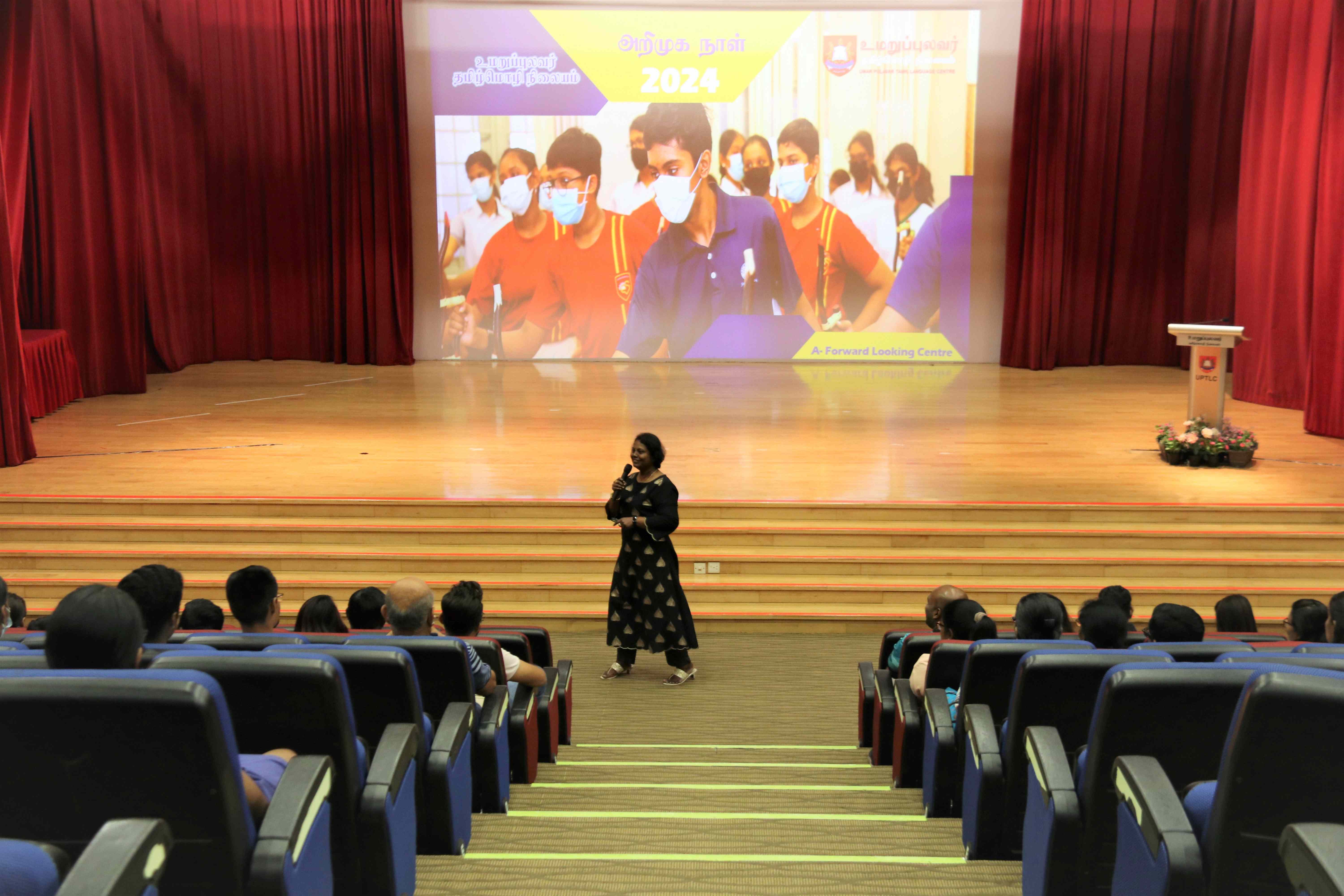 Woman speaks to seated audience in auditorium; large screen behind shows masked students and 2024 text.