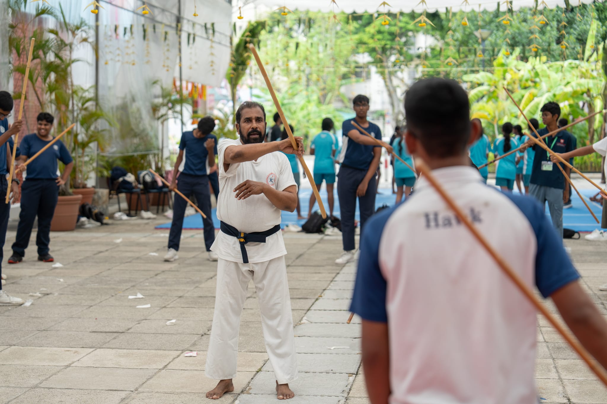 Man in white karate uniform demonstrates staff move outdoors as students practice with sticks behind him.