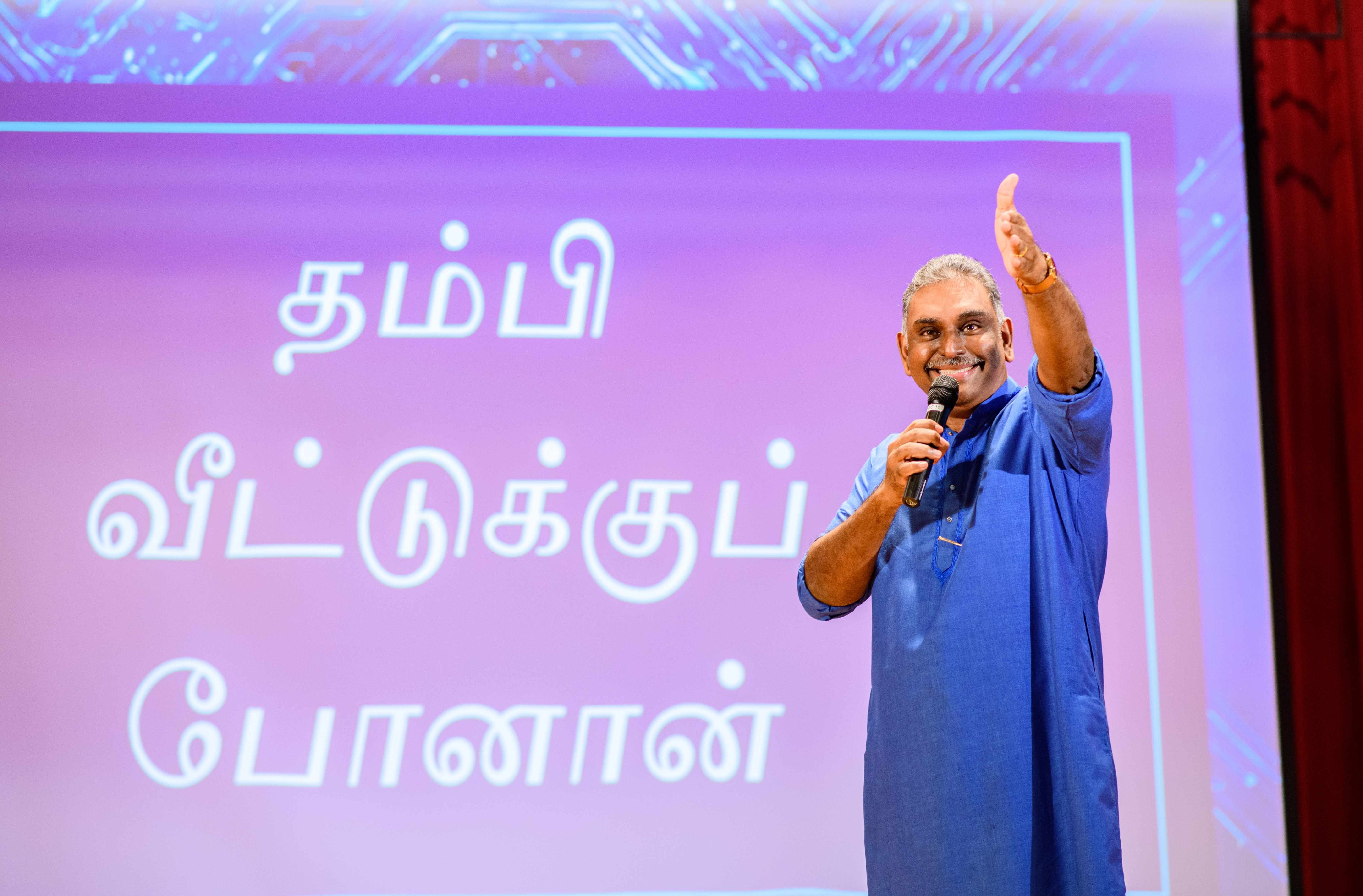 Man in blue shirt holds a microphone and waves on stage before a purple screen with Tamil text.