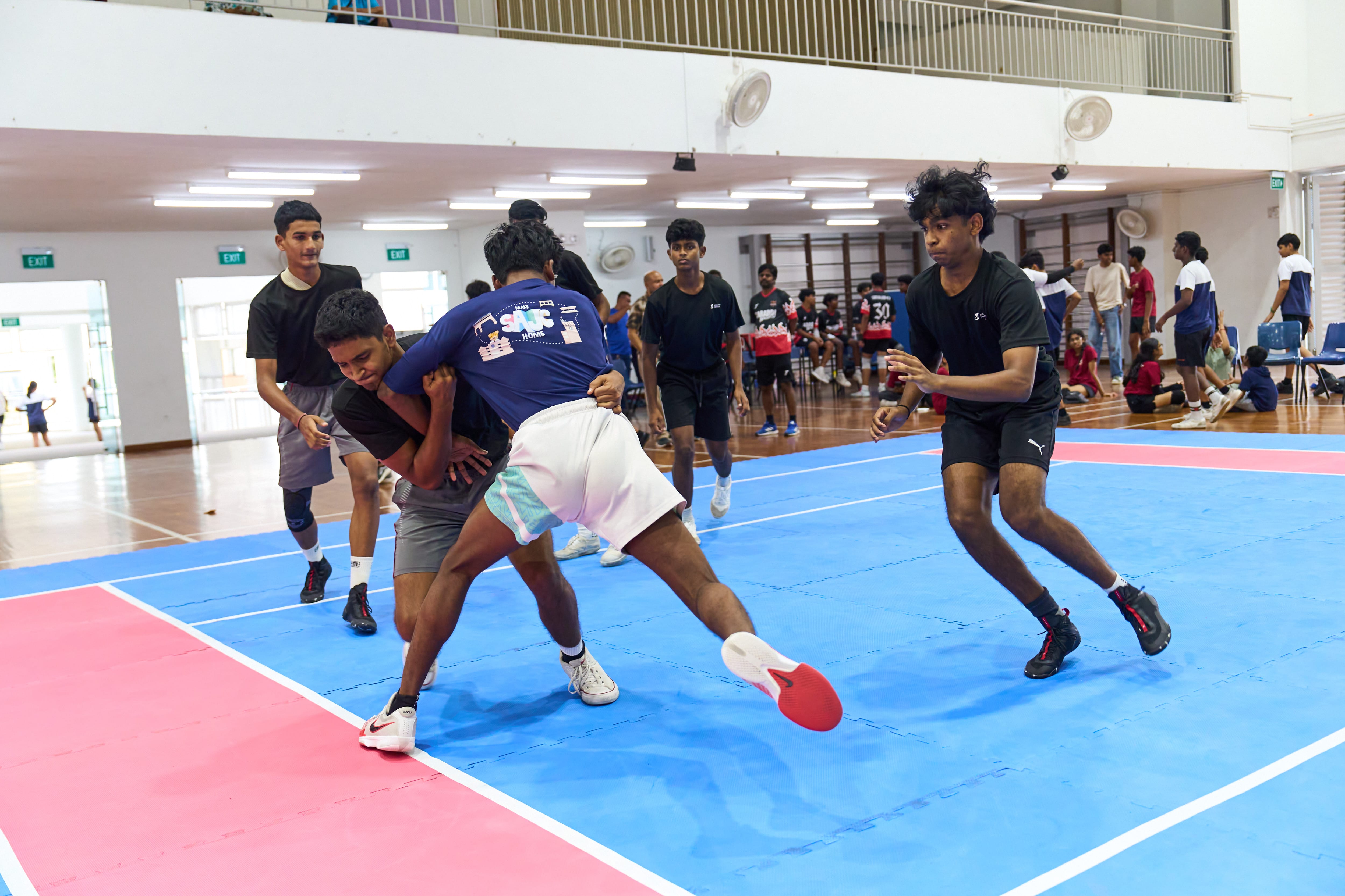 Two players grapple on a blue indoor court while another runs in; teammates and spectators watch behind.