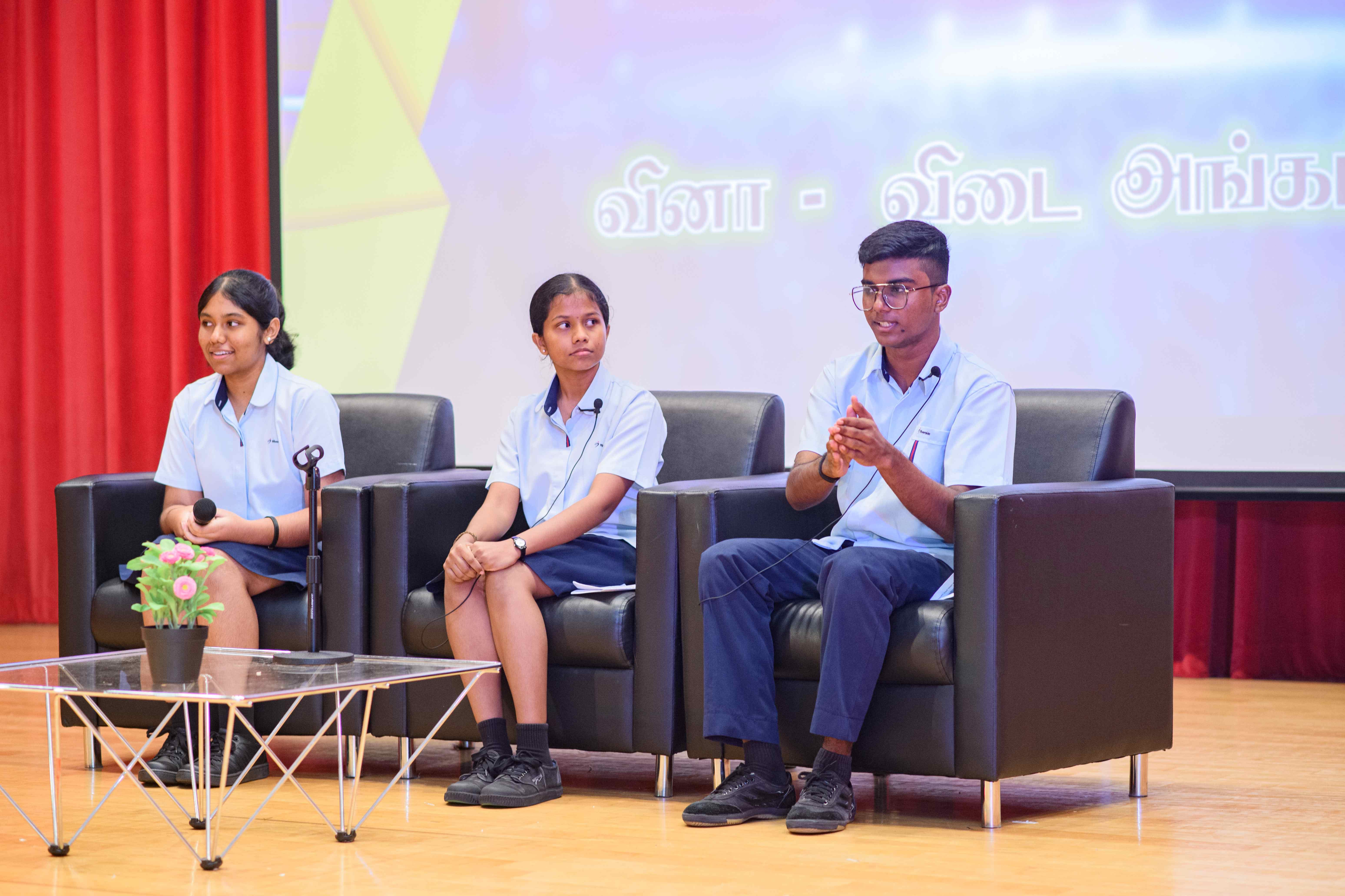 Three students in school uniforms sit on black armchairs onstage; one speaks as Tamil text shows behind.