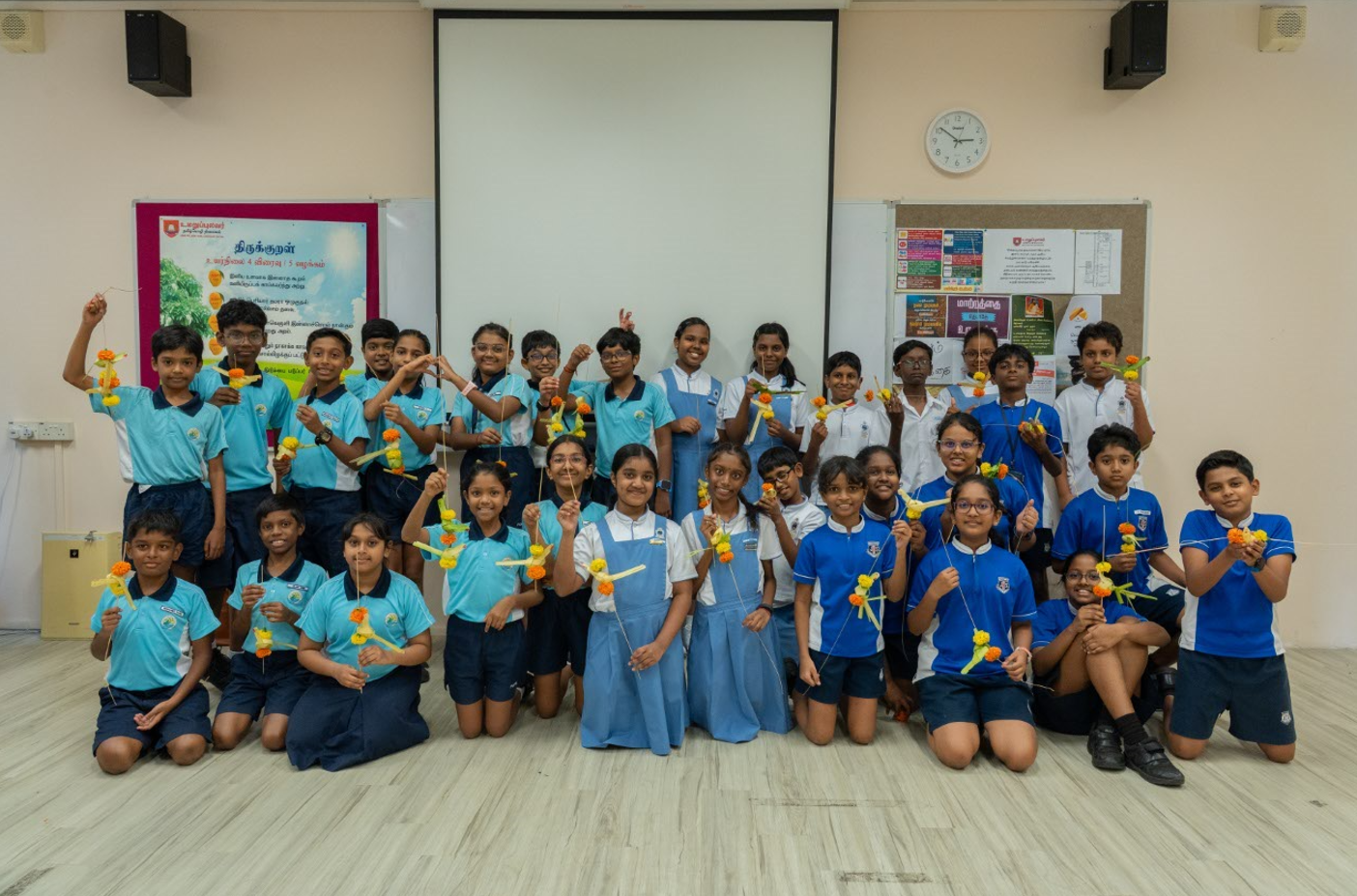 Group of schoolchildren in uniforms pose in a classroom, holding yellow and orange flower crafts.