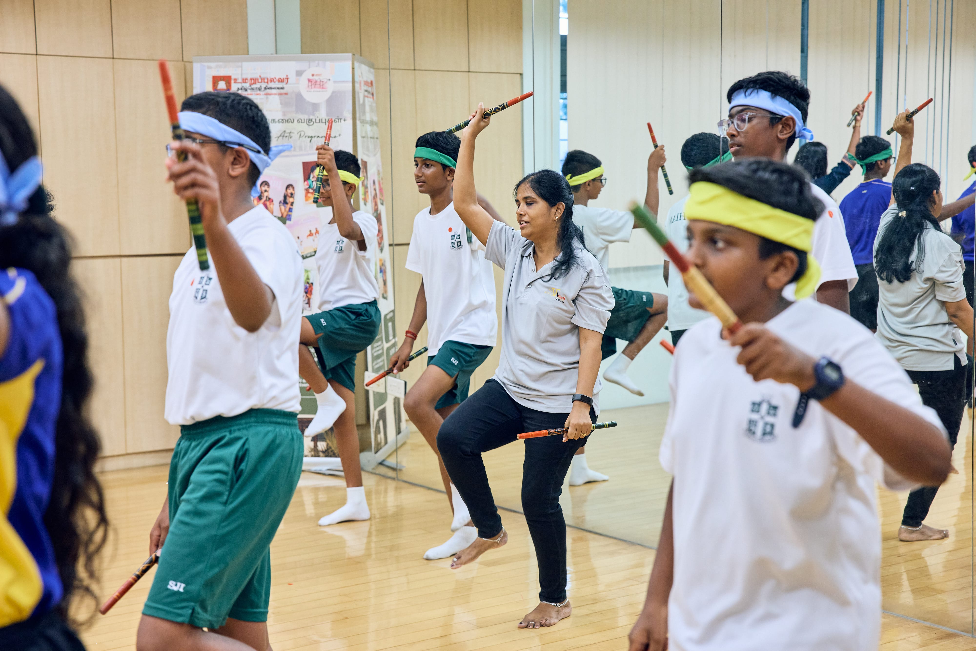 Students in a studio, wearing headbands, practicing with colorful sticks; instructor demonstrates a raised-knee pose.