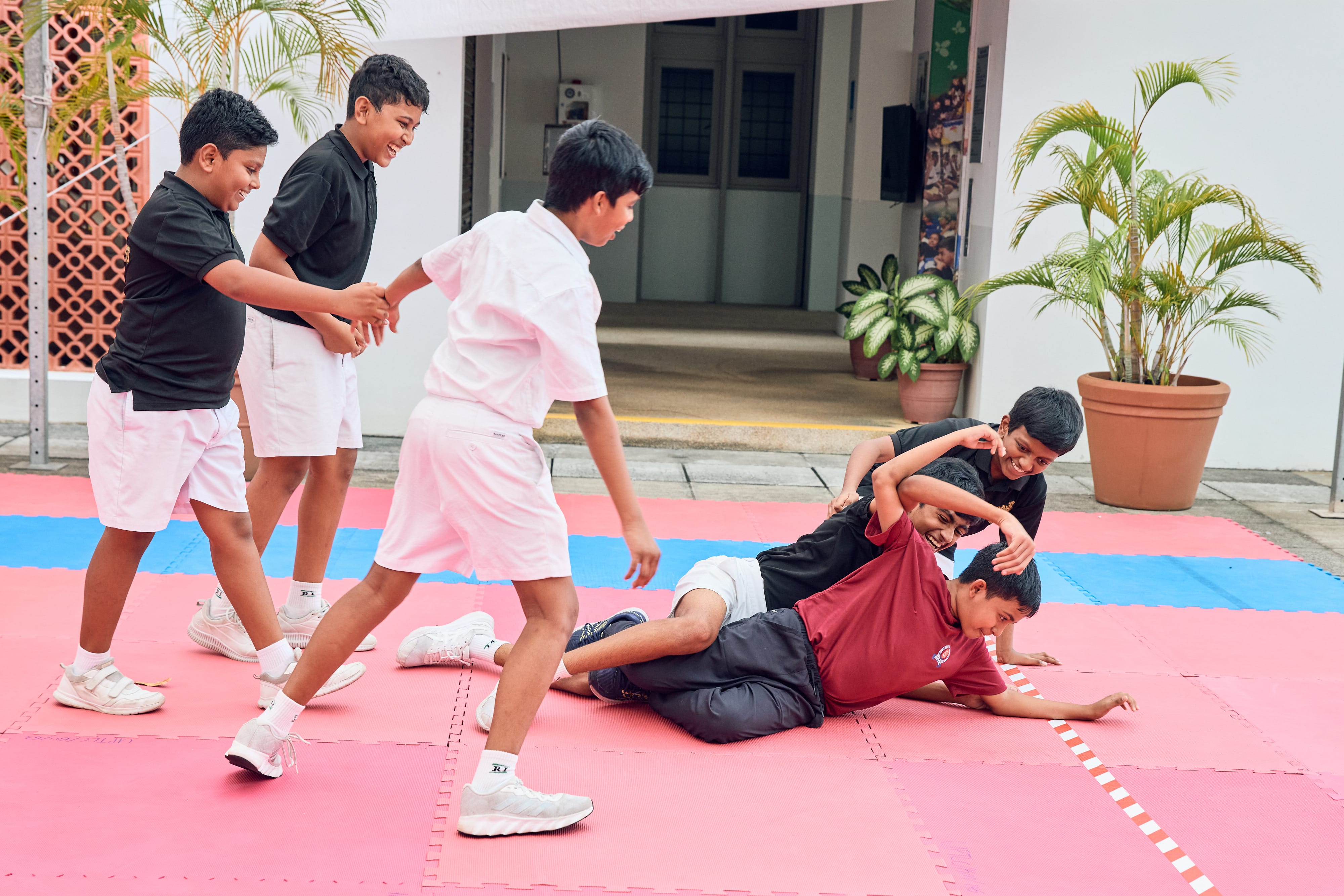 Five boys play wrestle on red and blue mats outside a building, with potted plants nearby.
