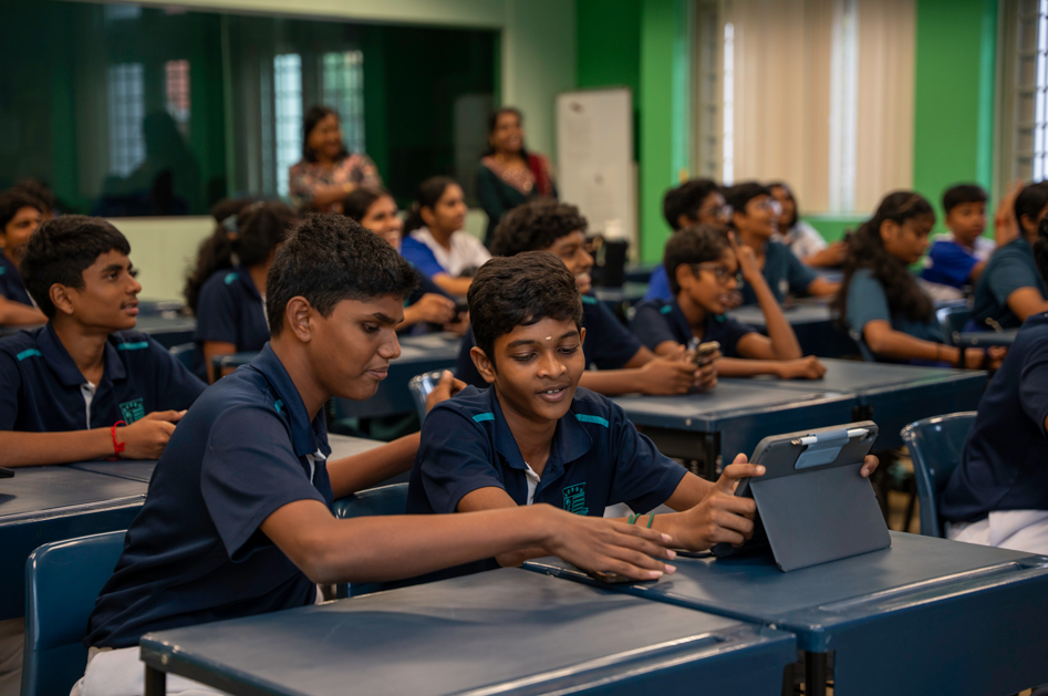 Two students in navy uniforms share a tablet at desks in a classroom with many classmates behind.