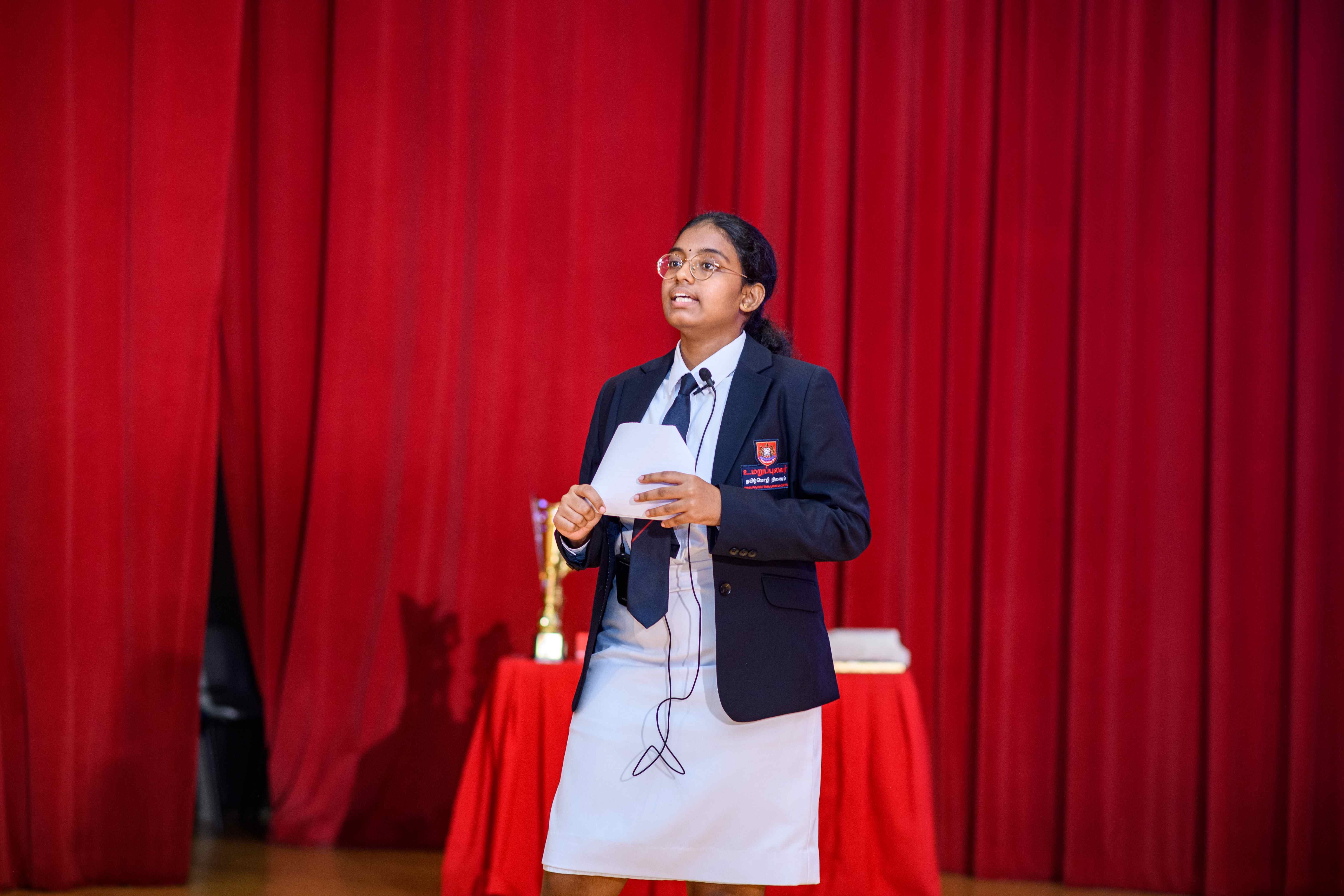 Student in blazer and tie speaks on stage before red curtains, holding notes with a mic wire visible.