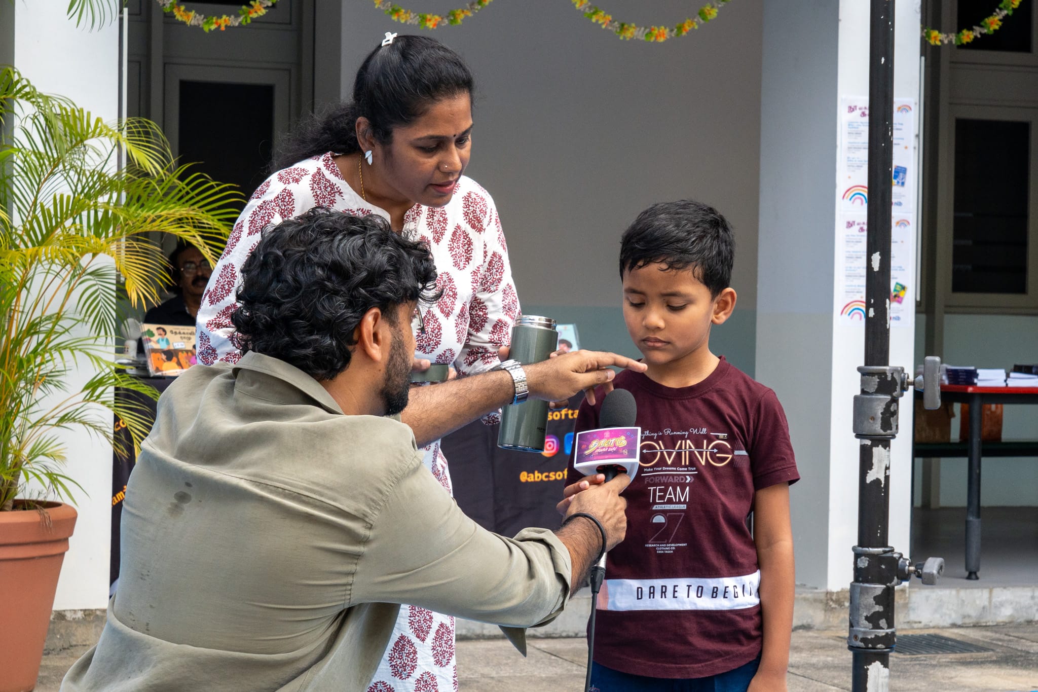 Man holds a microphone to a young boy as a woman stands behind them, outdoors near a building.