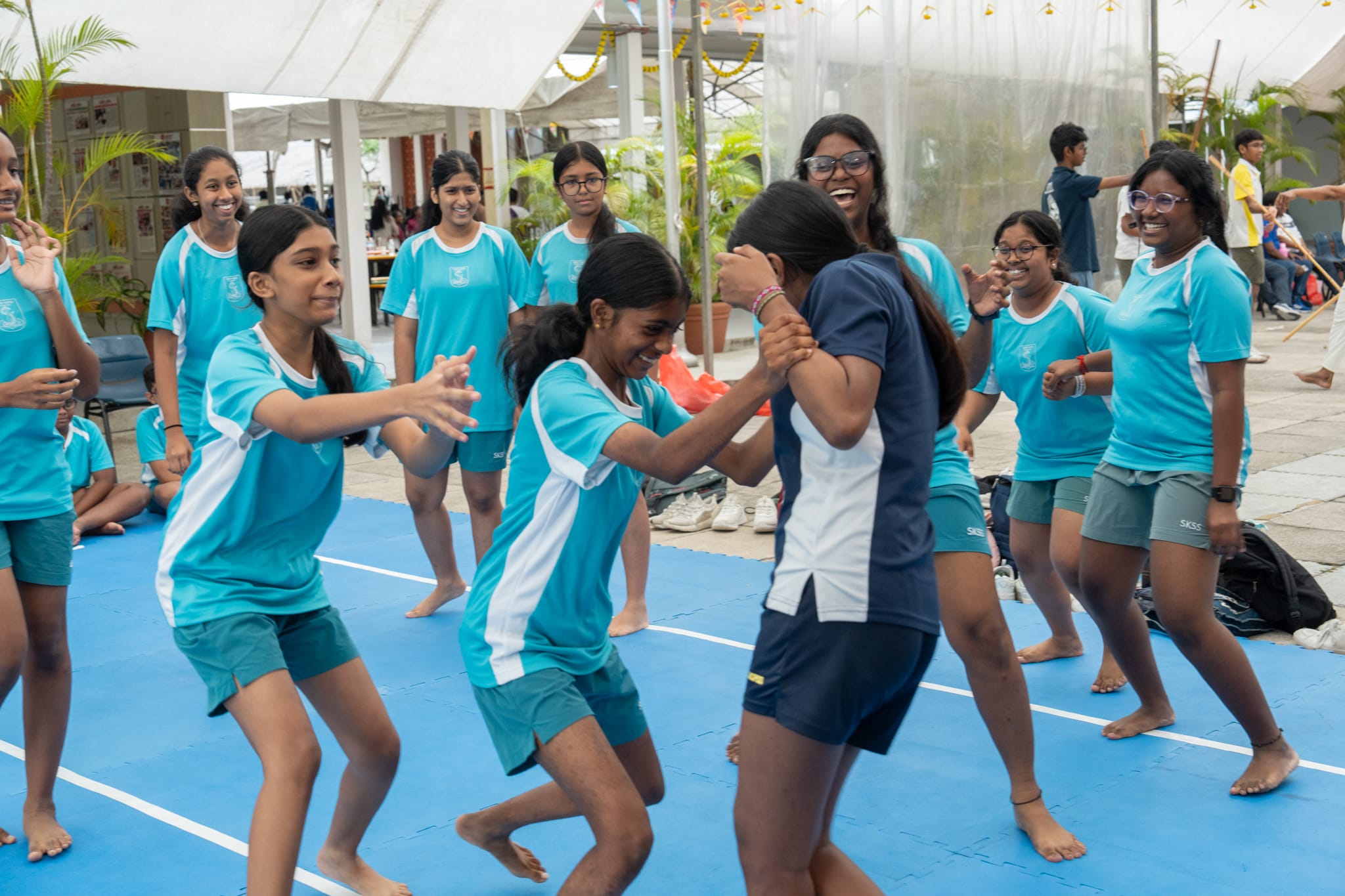 Group of girls in teal sports uniforms on a blue mat, laughing as two grapple in a game.