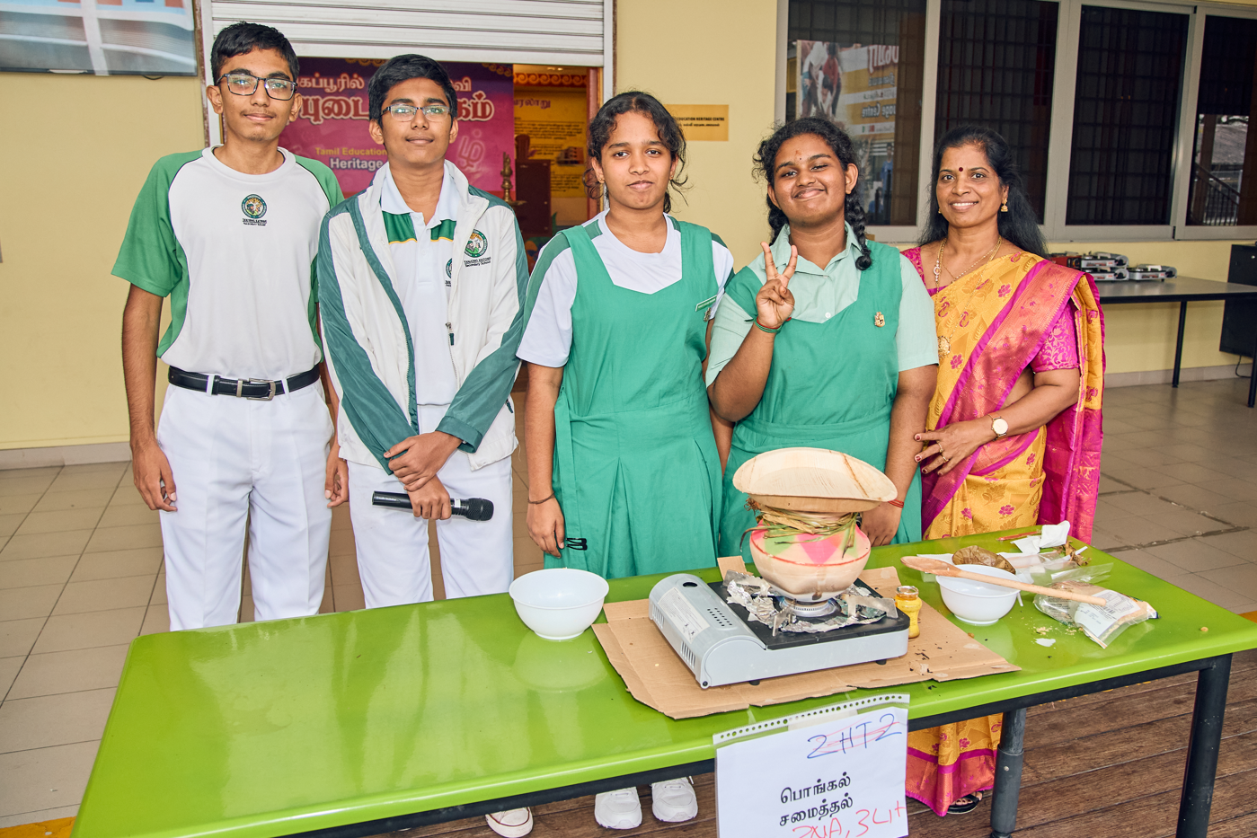 Five students and a teacher pose behind a table with a burner, pot, bowls, and food items.
