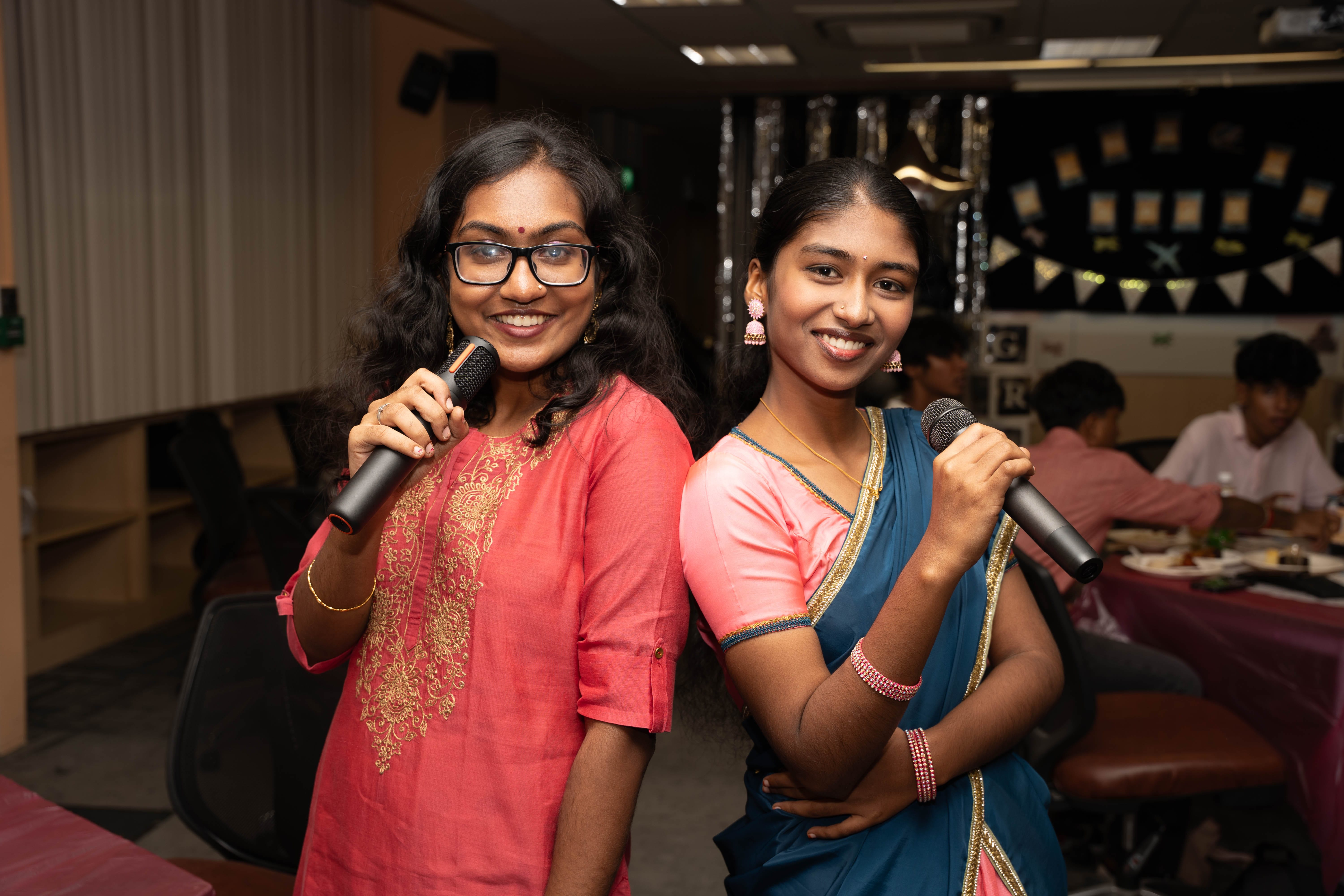 Two women smile holding microphones, one in a pink kurta and glasses, one in a blue sari at a party.