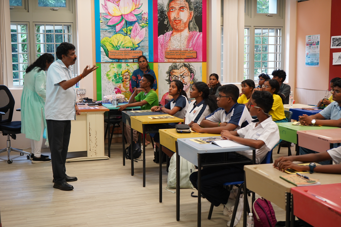 Teacher gestures while speaking to seated students in a classroom with colorful wall murals and tall windows.