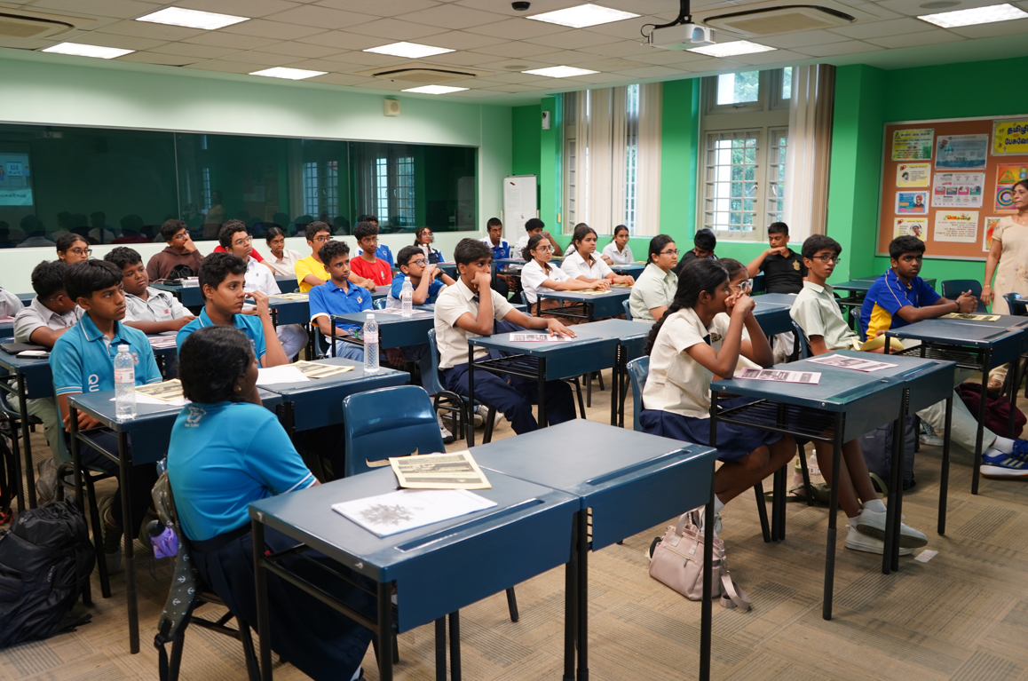 Classroom of seated students at blue desks facing a teacher near posters on a green wall.