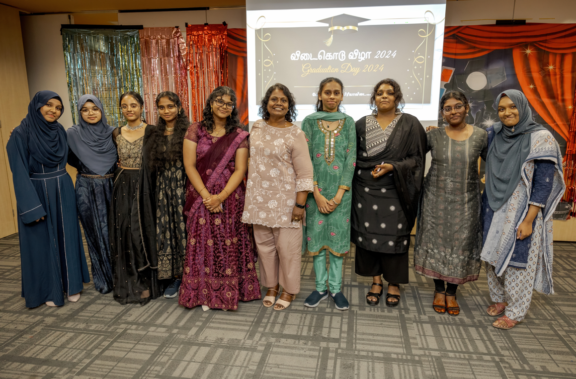 Nine women pose in formal attire on stage, with Graduation Day 2024 banner and shiny curtains behind.