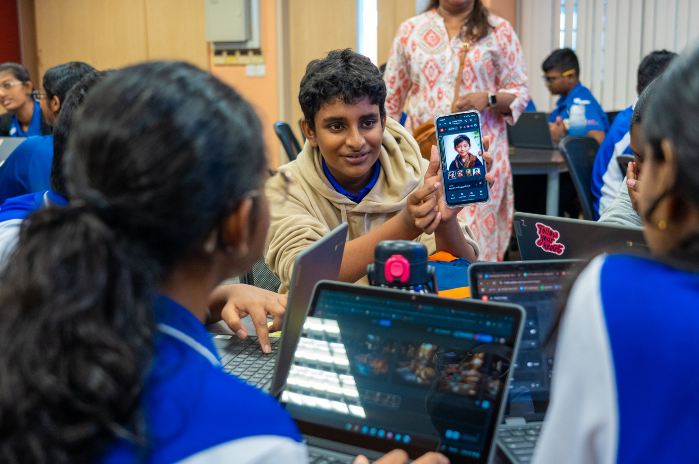 Student in beige hoodie shows a phone screen to classmates seated with open laptops in a classroom.