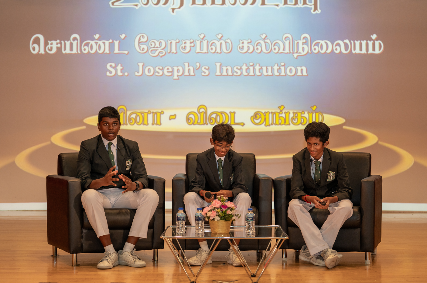 Three students in school uniforms sit on armchairs on stage with a table, flowers and water bottles behind text.