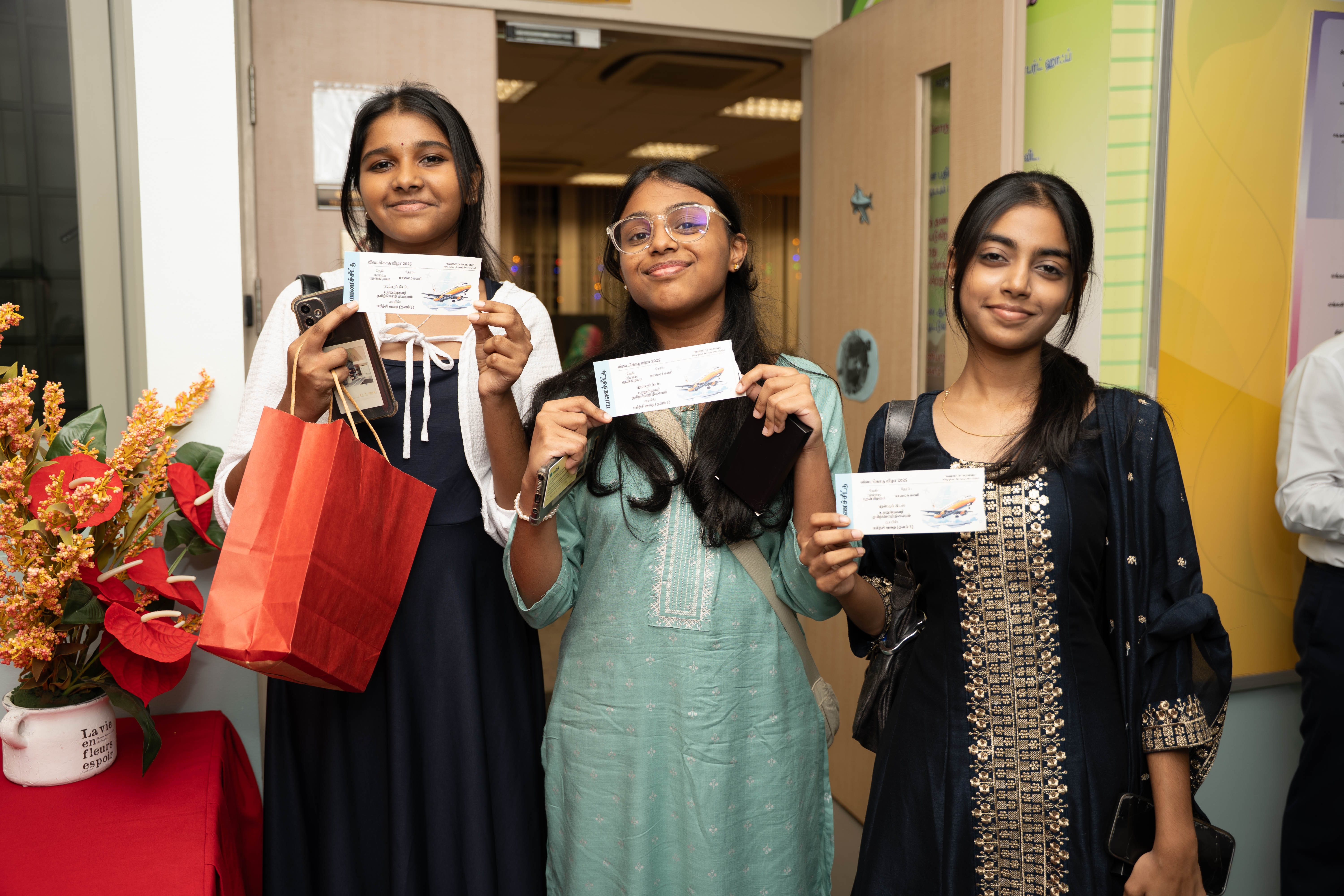 Three young women smile indoors, each holding a paper ticket; one holds a red gift bag.