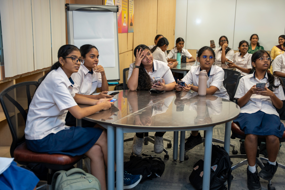 Students in school uniforms sit around a round table, some holding phones, watching something off-camera.