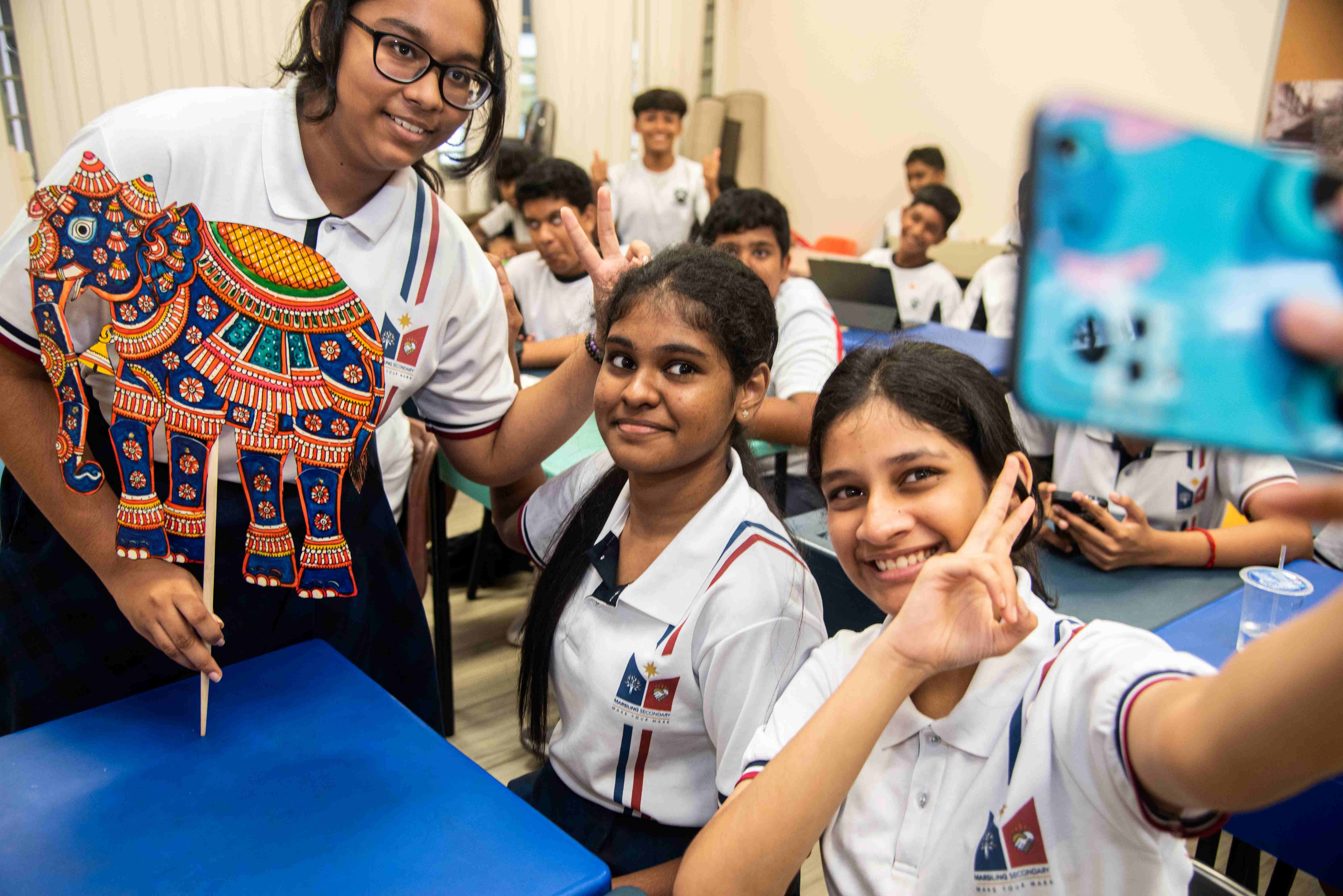 Students in uniforms smile at a classroom selfie; one holds a colorful elephant cutout on a stick.