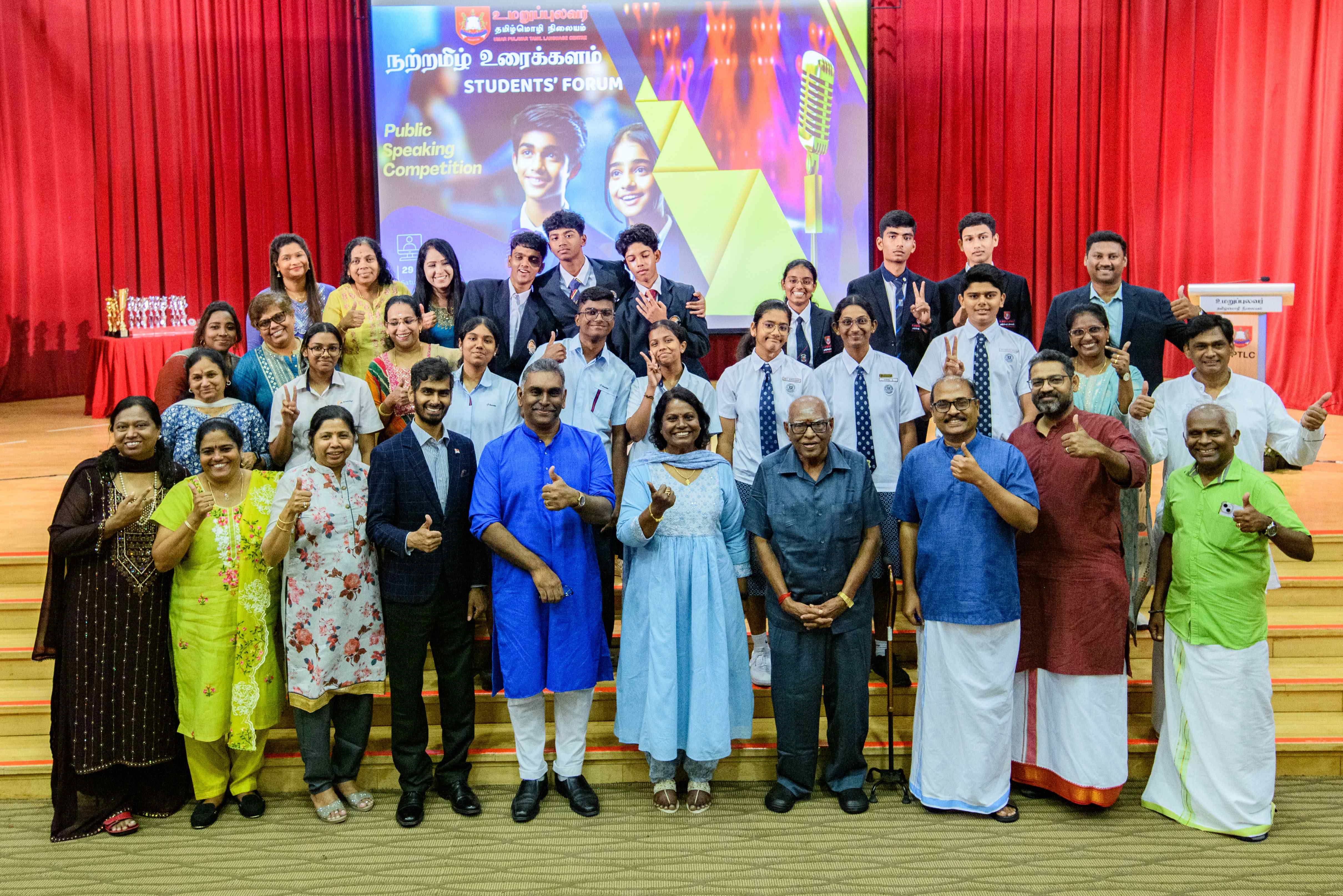 Group of students and adults posing on stage before a Students' Forum public speaking backdrop, many thumbs up.