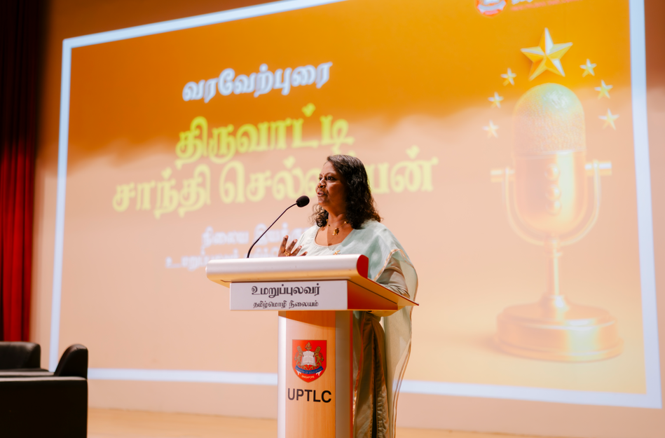 Woman in sari speaks at a podium with microphone; orange backdrop shows Tamil text and a gold mic icon.