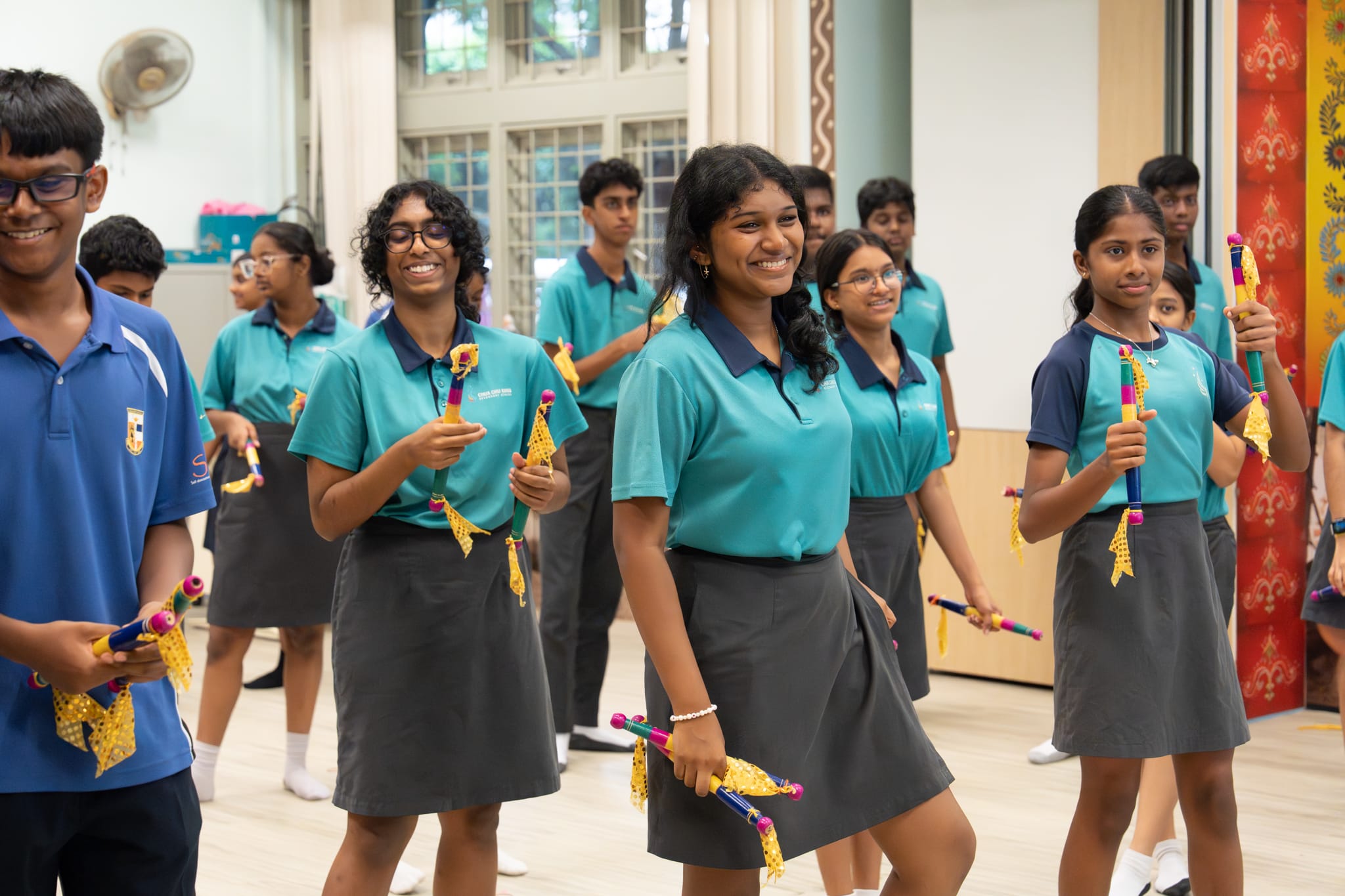 Smiling students in teal uniforms hold colorful decorated sticks in a classroom, as if practicing a dance.