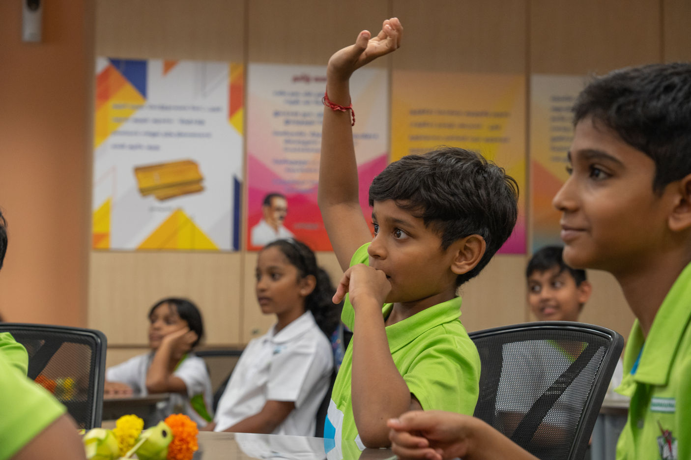 Boy in green shirt raises his hand in a classroom while other children sit behind him.