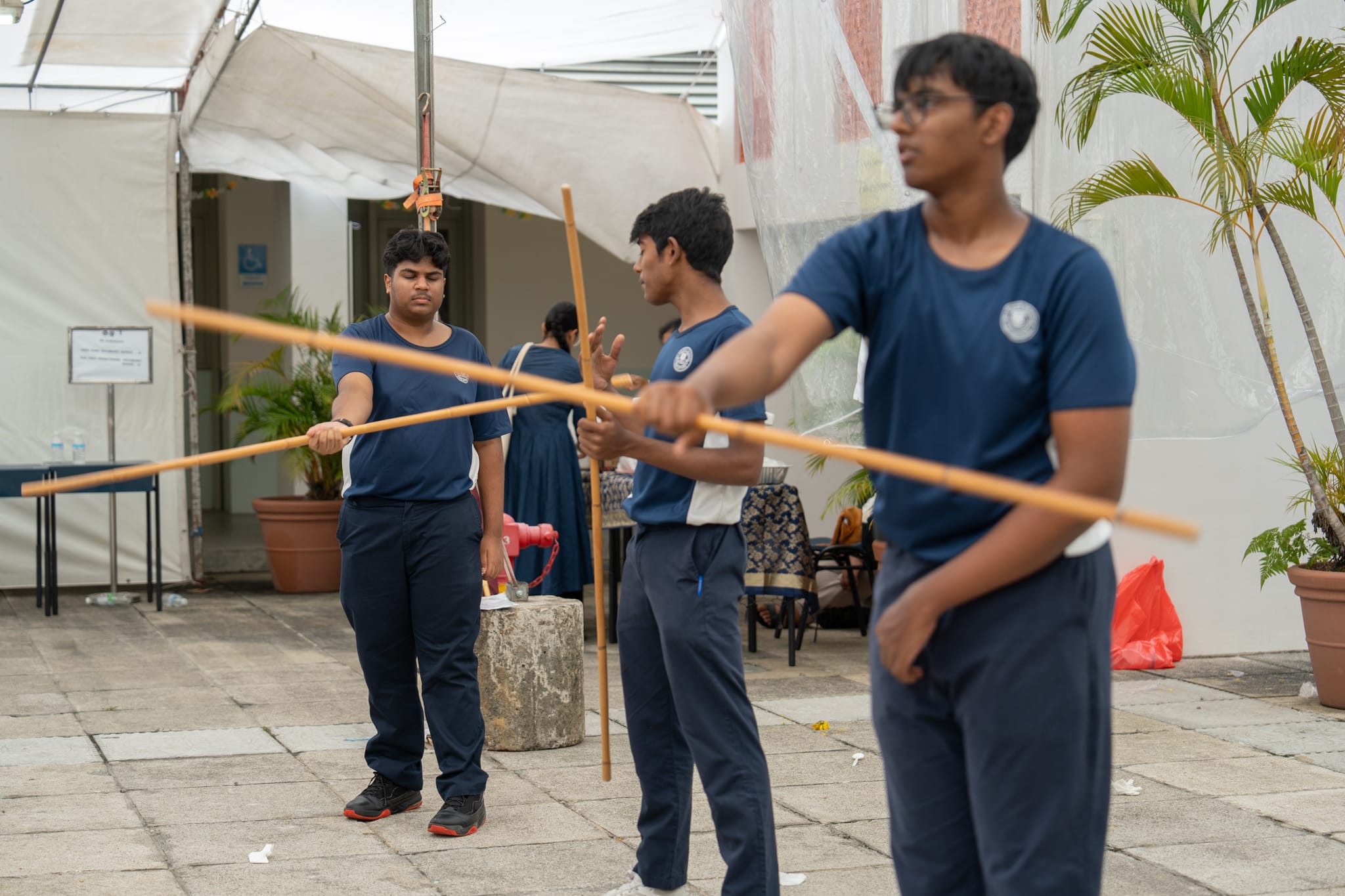 Three youths in blue shirts practice staff moves outdoors under a canopy, with potted plants nearby.