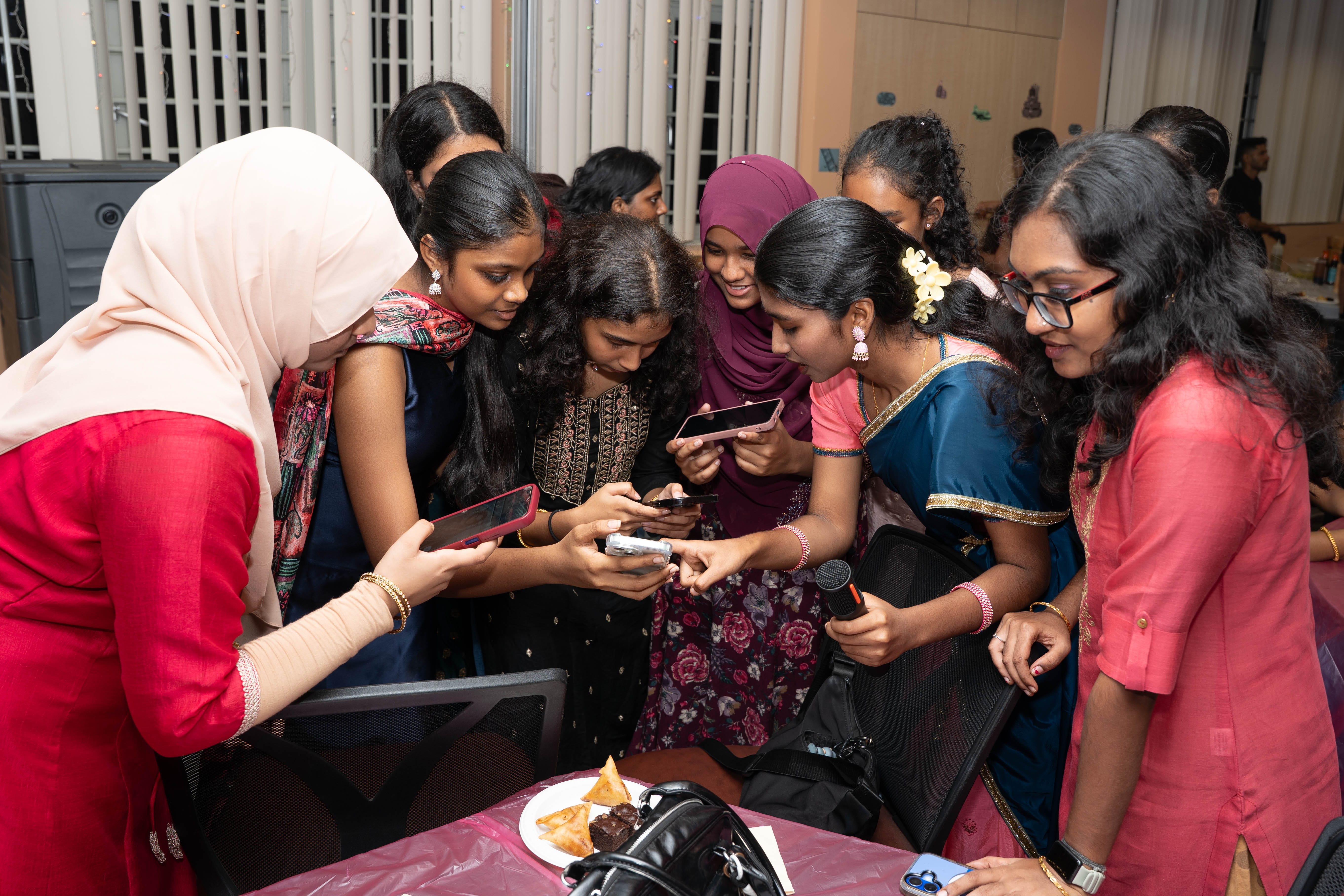 Group of women crowd around a table, looking at phones; one holds a microphone near snacks and bags.