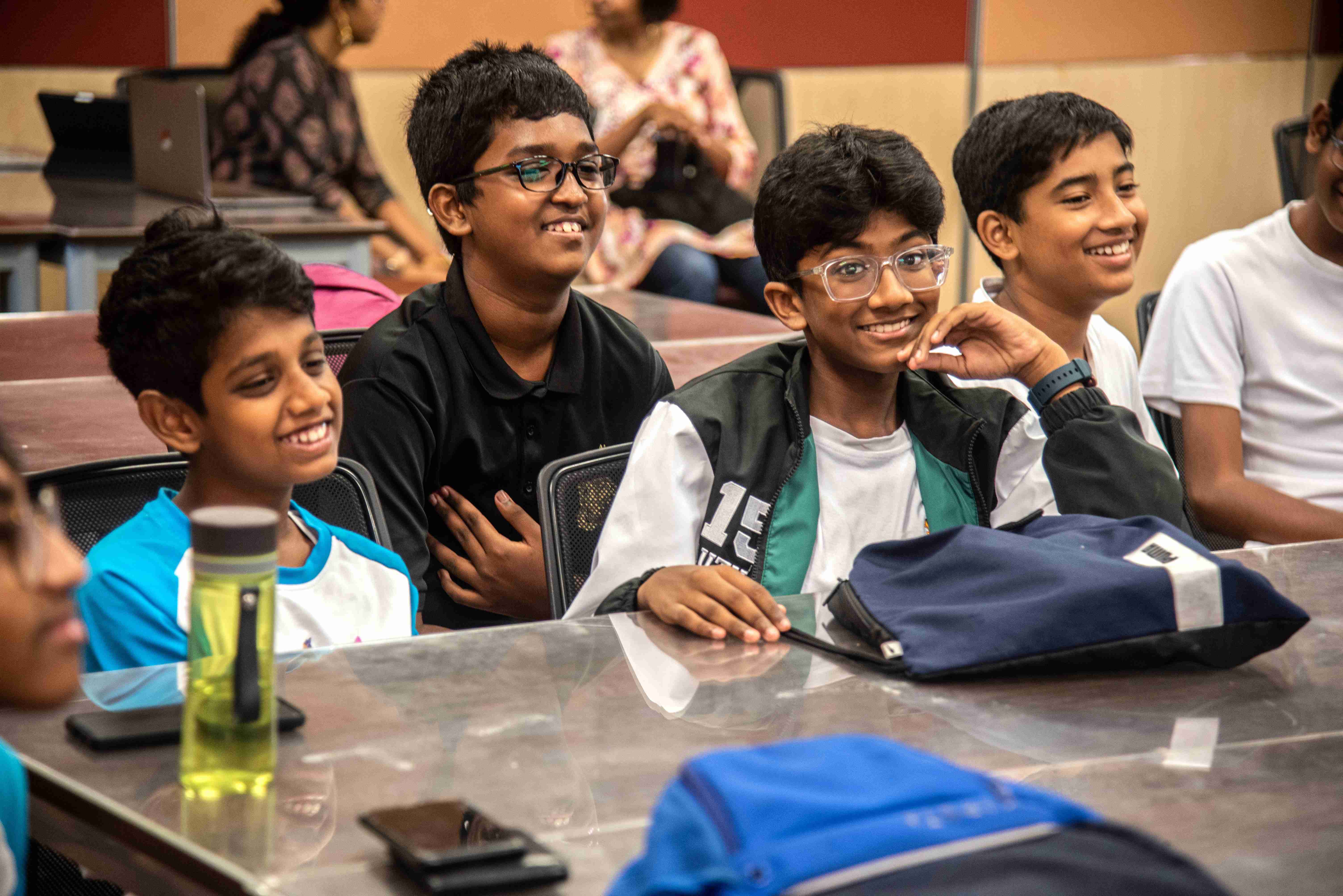 Four boys smiling at a classroom table; two wear glasses, with backpacks and a water bottle in front.