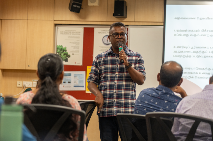 Man in plaid shirt speaks into a microphone in a classroom, with seated attendees and a projected slide.