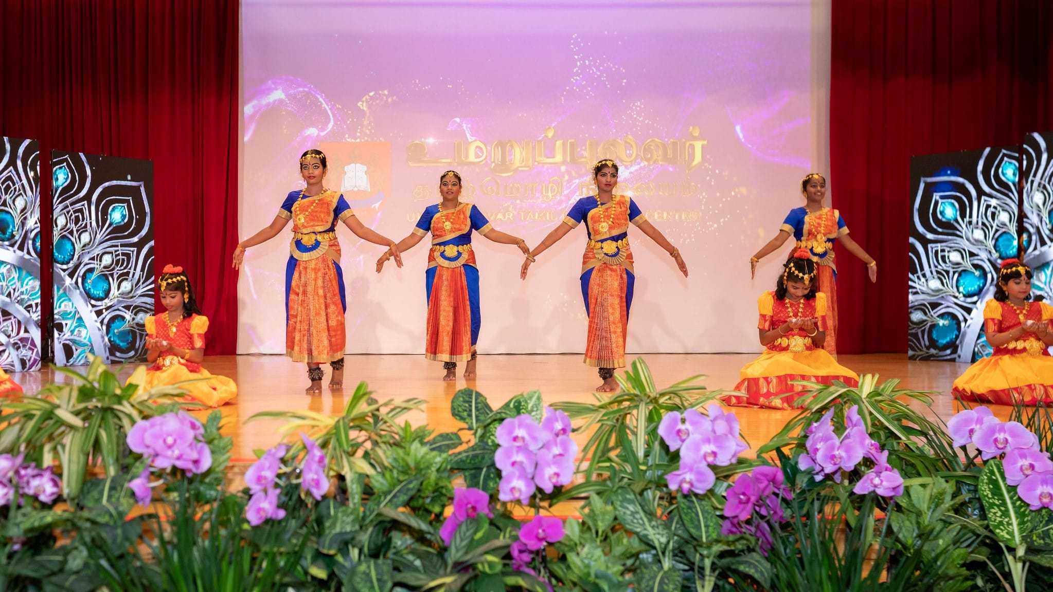 Six dancers in bright traditional costumes perform on a stage with red curtains and purple flowers in front.