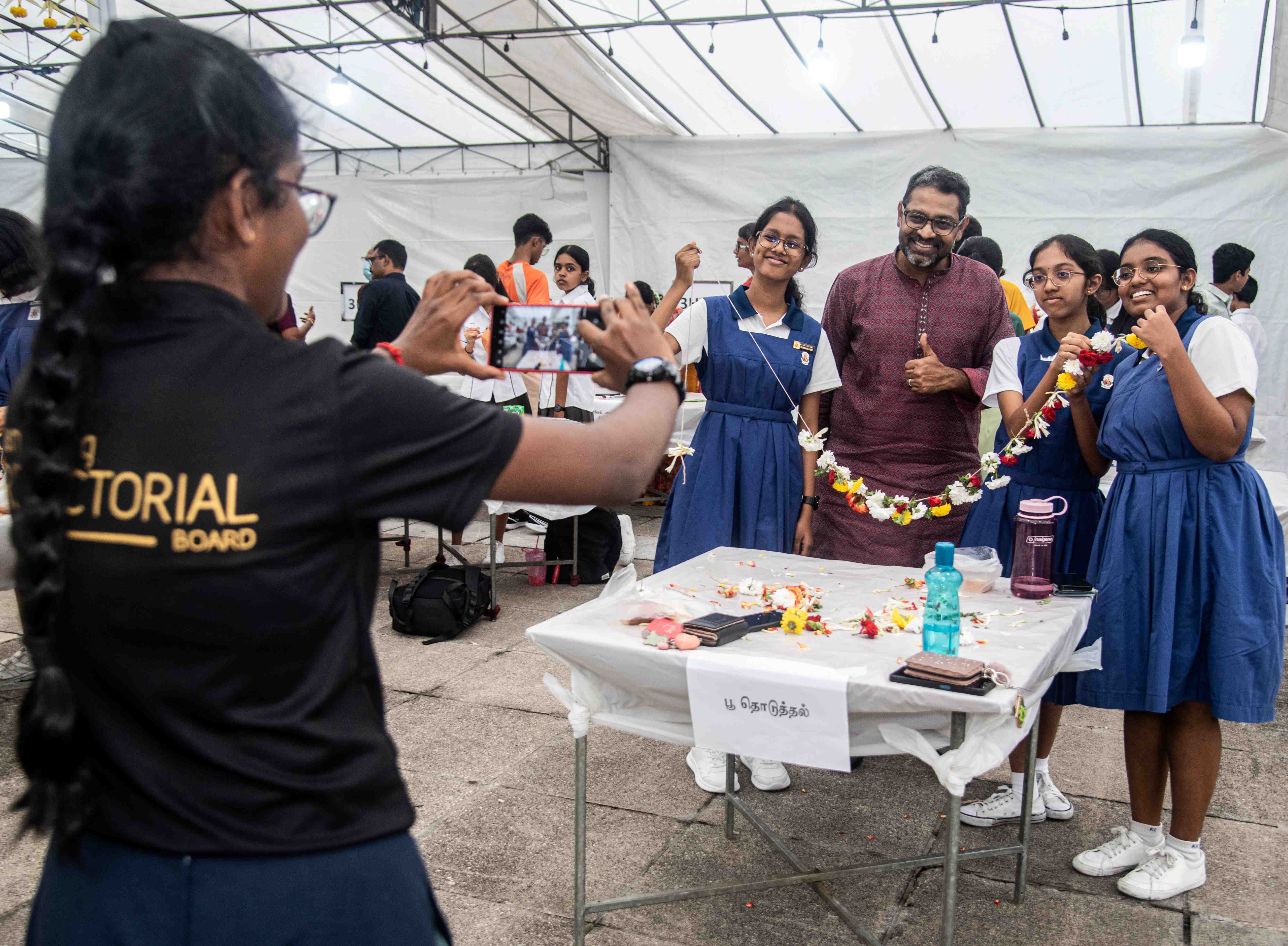Girl photographs three students posing with a man; they hold a flower garland behind a table.