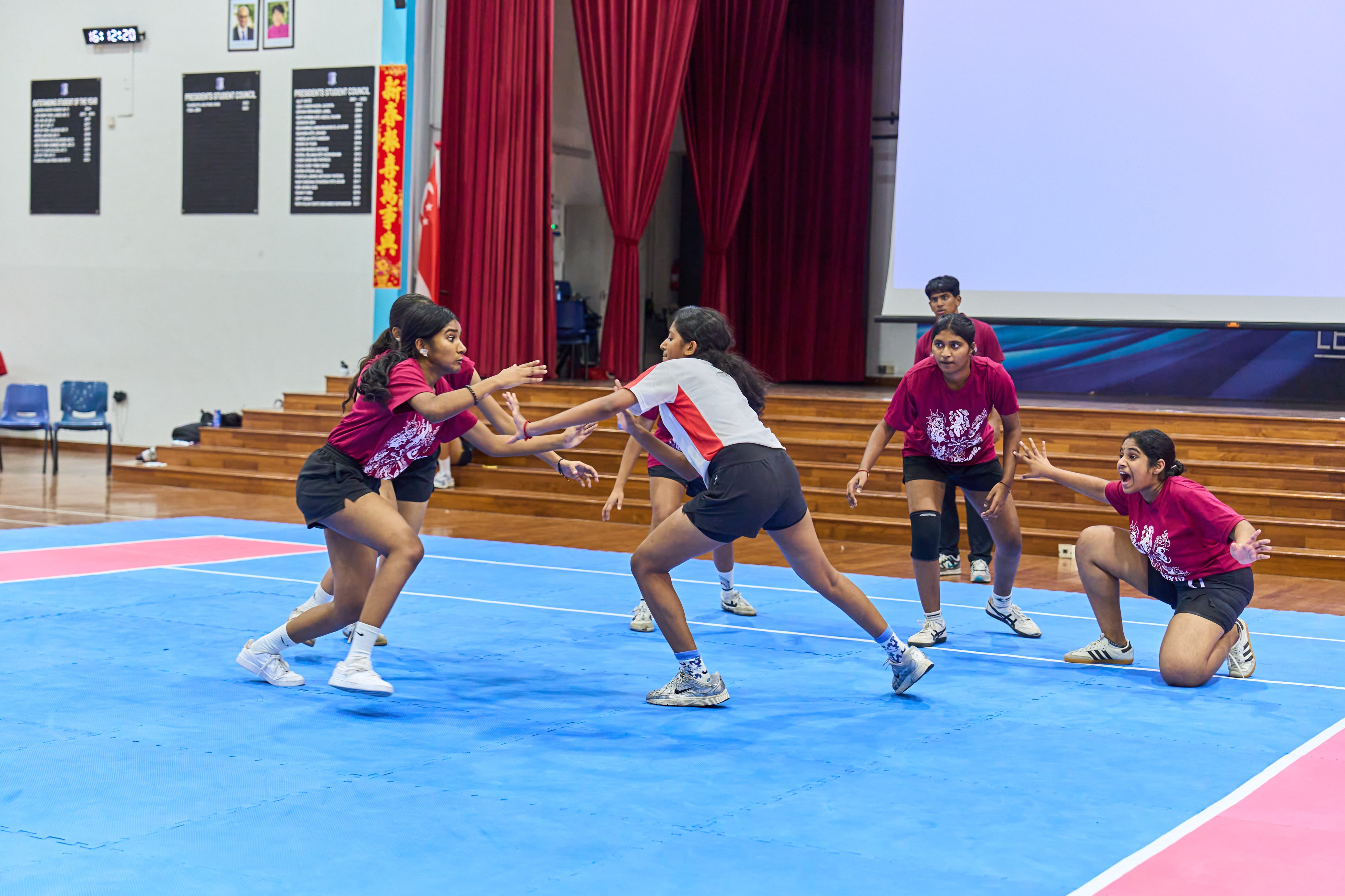 Girls in sports jerseys play kabaddi on a blue mat indoors, reaching and crouching to tag opponents.