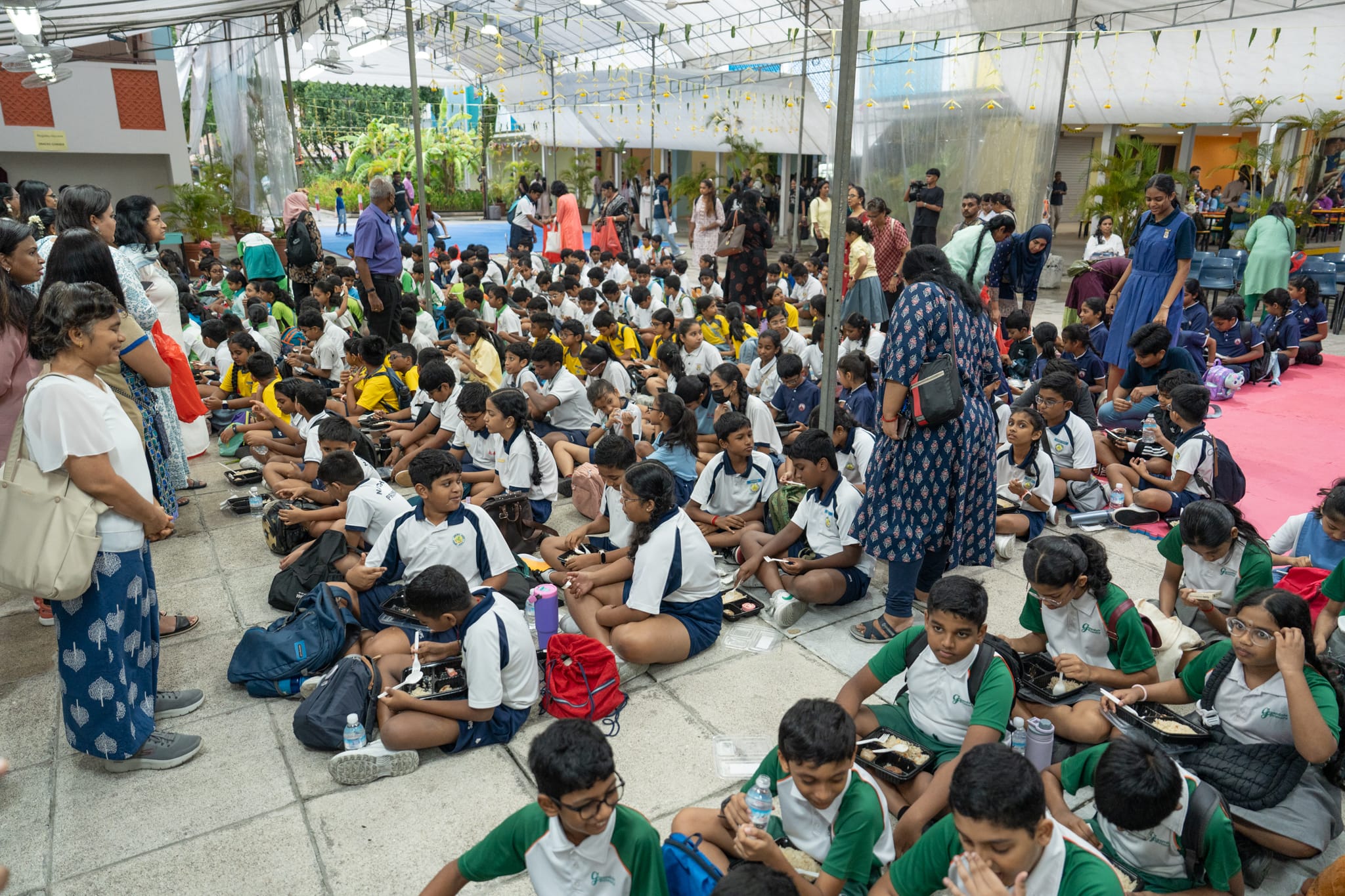 Crowd of students in uniforms sit on the ground eating lunch under a covered courtyard while adults stand nearby.