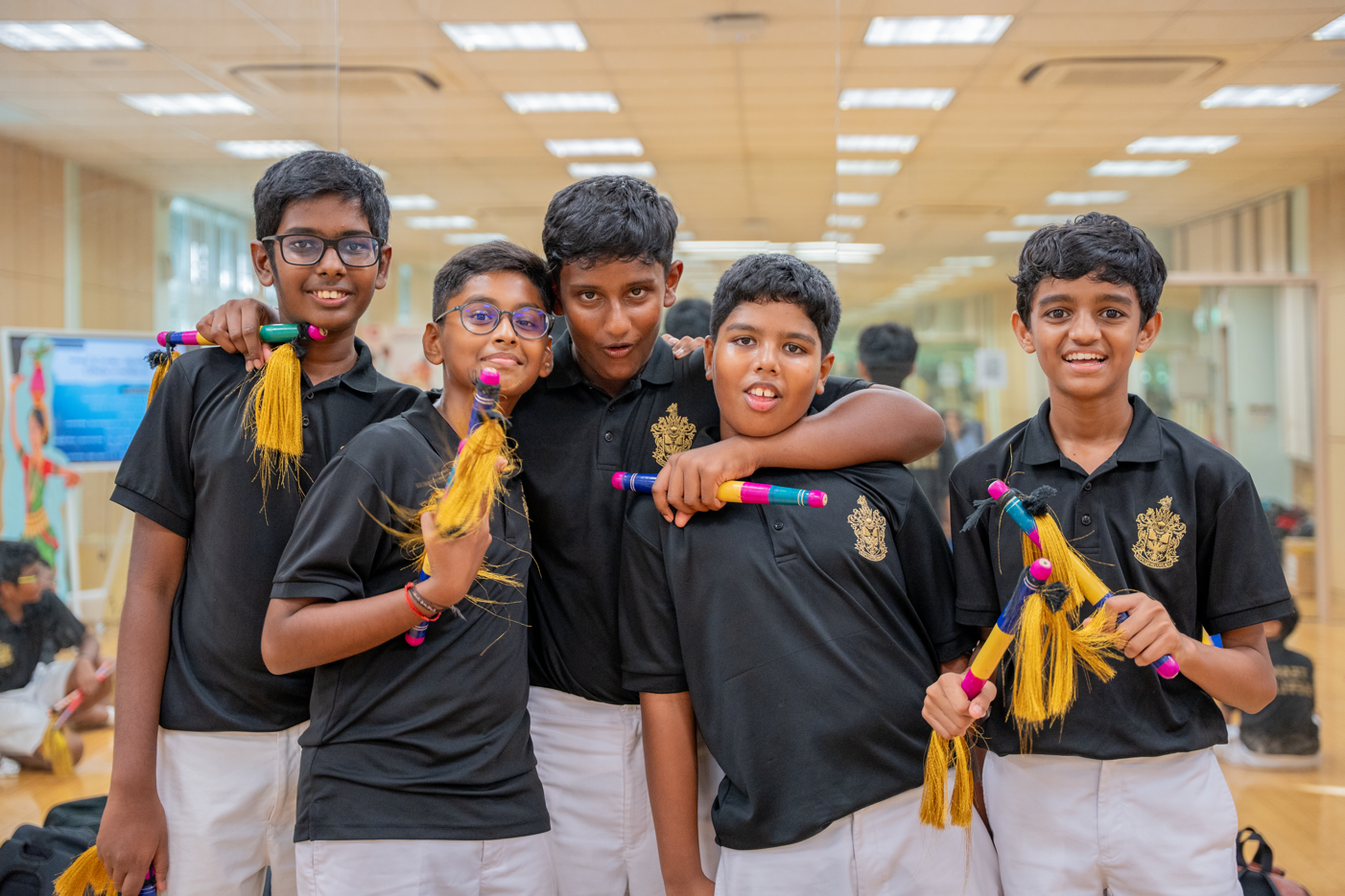 Five boys in black polos and white pants pose indoors, smiling and holding colorful tasselled sticks.