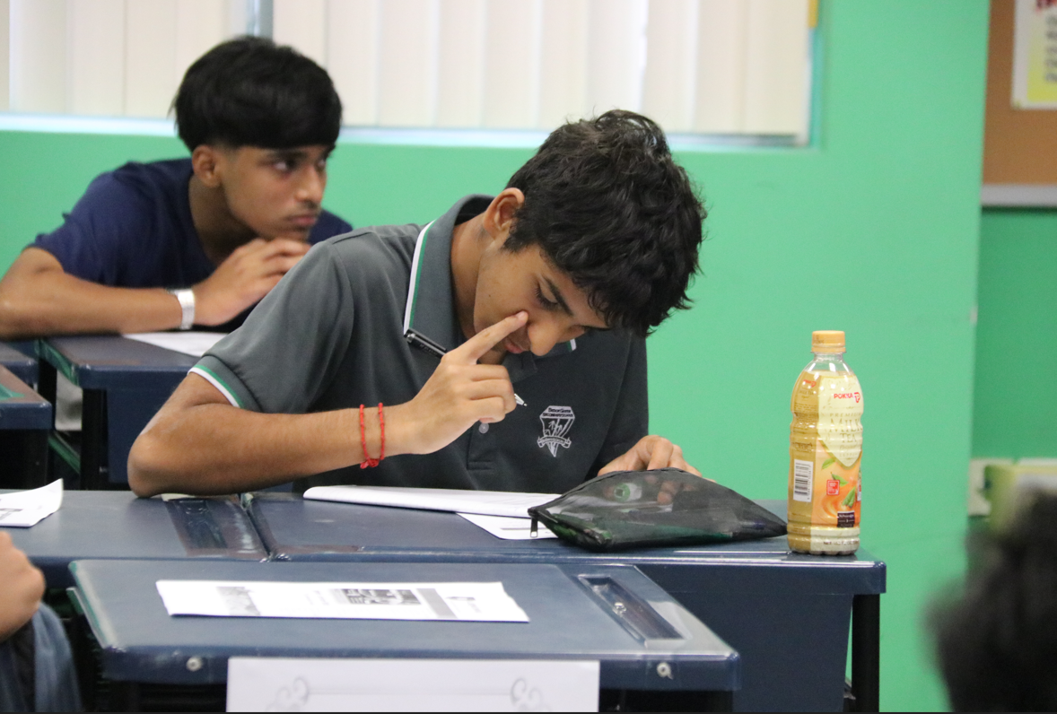 Student taking a test at a desk, finger on nose, with another student behind and a drink bottle nearby