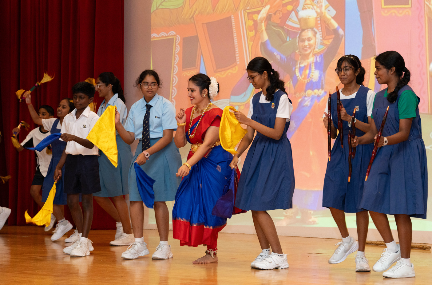 Schoolchildren on stage dance with colorful scarves; a woman in red and blue sari dances center.