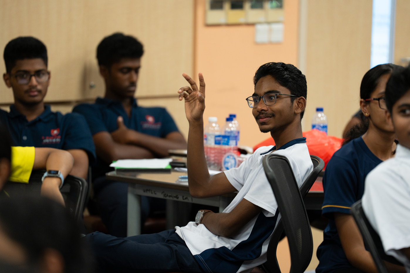 Seated student in glasses raises a hand in a classroom, with other students and water bottles behind.
