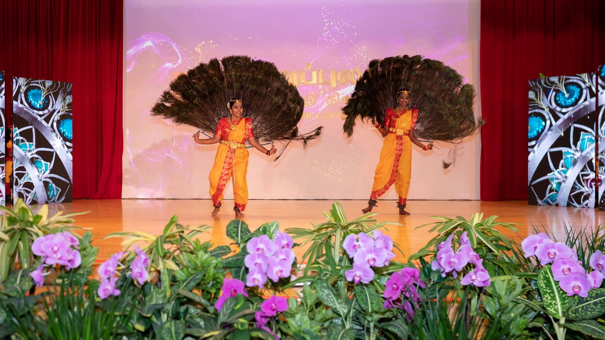 Two dancers in yellow and red costumes perform on stage holding large peacock-feather fans; flowers in front.